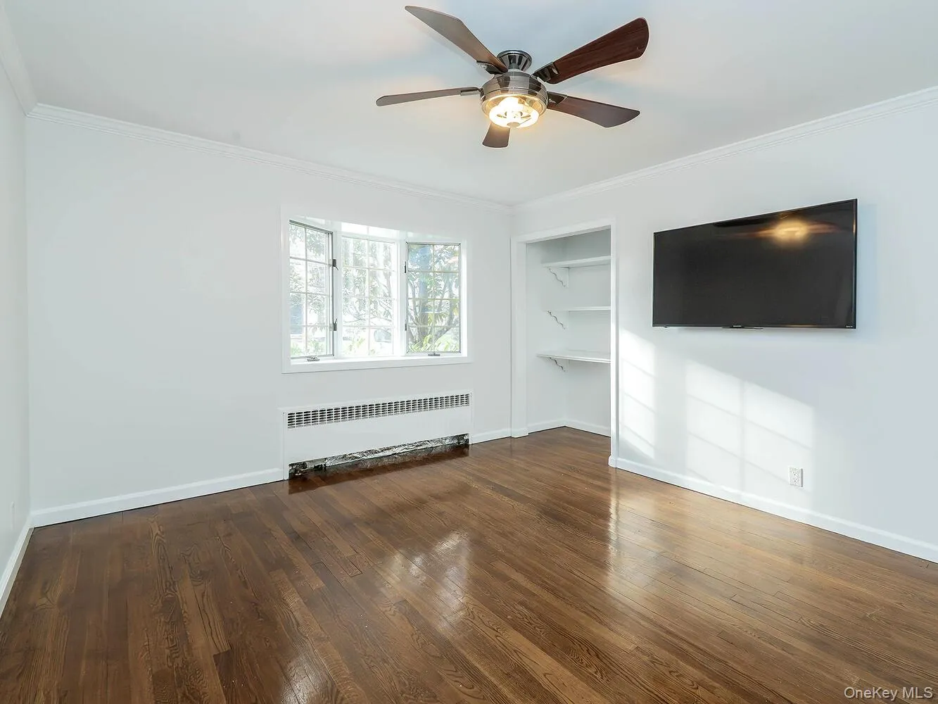 Unfurnished living room featuring crown molding, radiator, built in shelves, dark wood-type flooring, and ceiling fan Unfurnished living room featuring crown molding, radiator, built in shelves, dark wood-type flooring, and ceiling fan