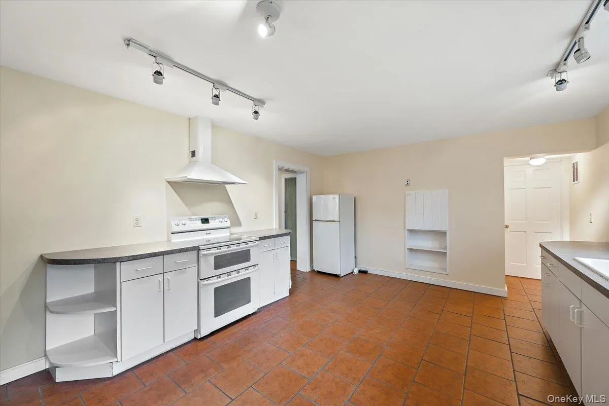 Kitchen with rail lighting, white appliances, wall chimney exhaust hood, open shelves, and white cabinets Kitchen with rail lighting, white appliances, wall chimney exhaust hood, open shelves, and white cabinets