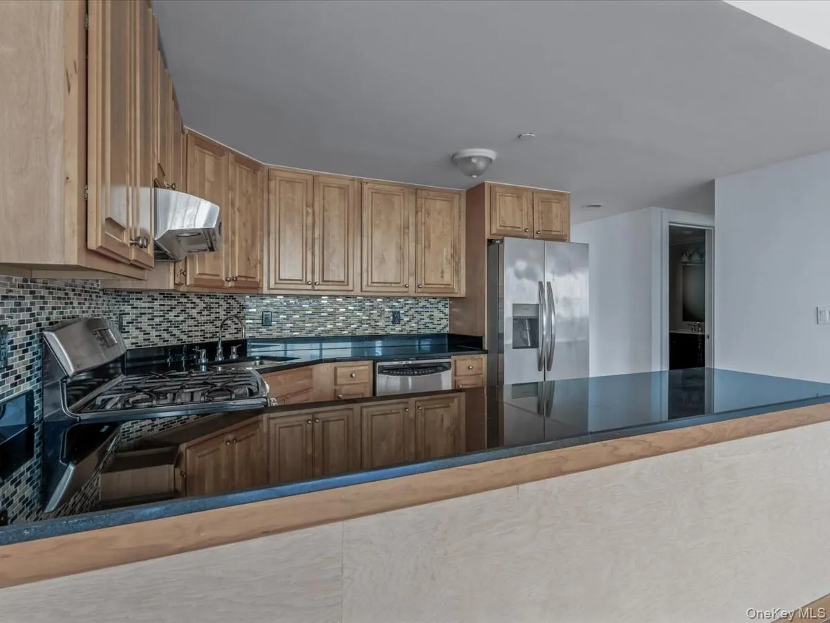 Kitchen featuring appliances with stainless steel finishes, tasteful backsplash, brown cabinetry, under cabinet range hood, and dark stone countertops Kitchen featuring appliances with stainless steel finishes, tasteful backsplash, brown cabinetry, under cabinet range hood, and dark stone countertops