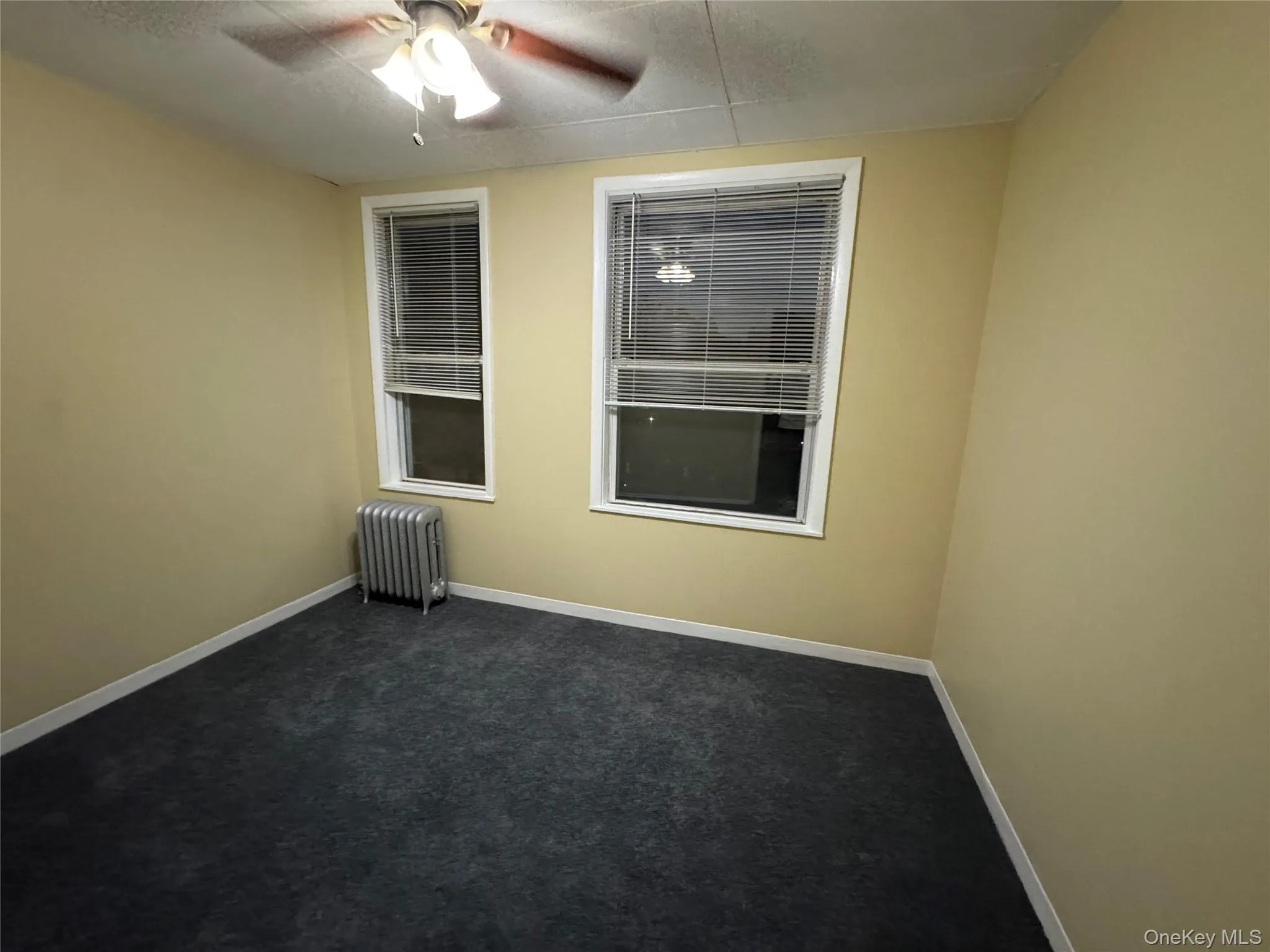 Unfurnished room featuring radiator heating unit, dark colored carpet, and ceiling fan Unfurnished room featuring radiator heating unit, dark colored carpet, and ceiling fan