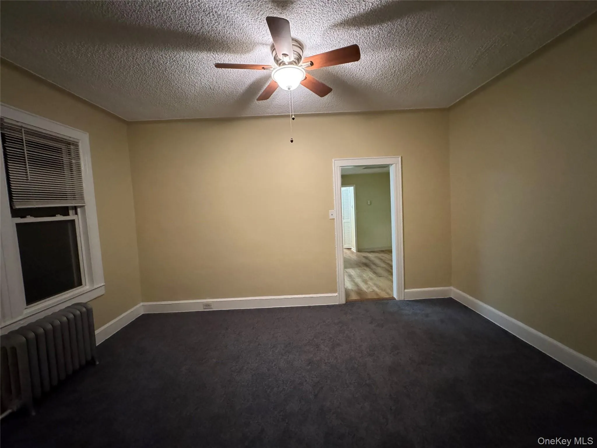 Unfurnished room with radiator, dark colored carpet, a textured ceiling, and a ceiling fan Unfurnished room with radiator, dark colored carpet, a textured ceiling, and a ceiling fan