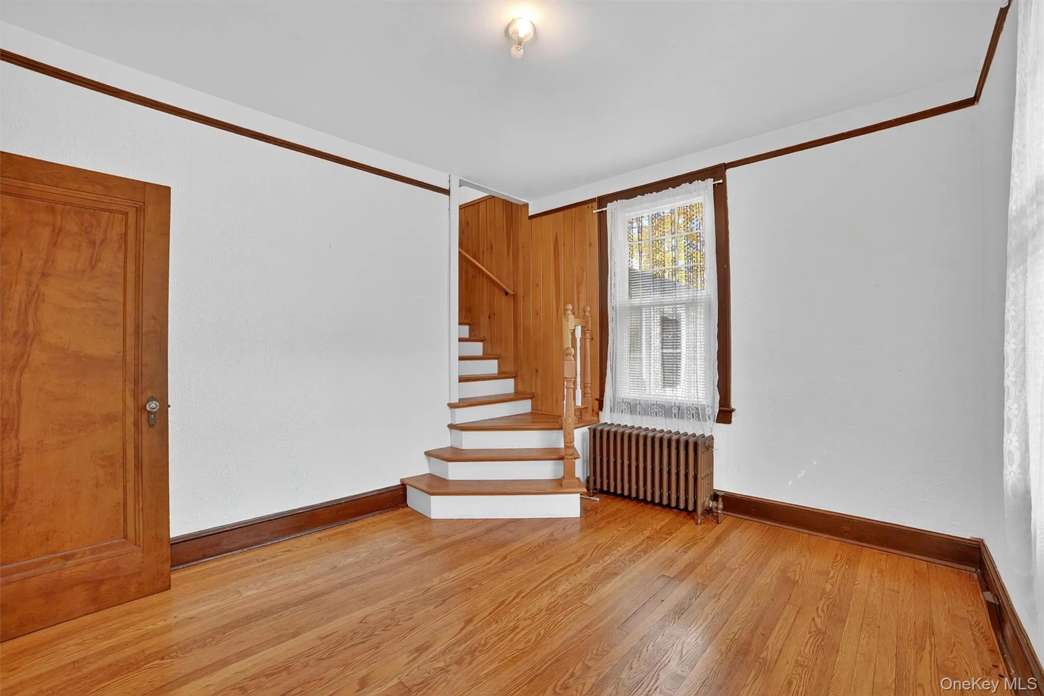 Spare room with light wood-type flooring, radiator, stairway, ornamental molding, and wooden walls Spare room with light wood-type flooring, radiator, stairway, ornamental molding, and wooden walls