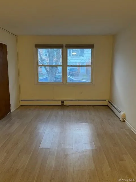 Empty room featuring healthy amount of natural light, light wood-style flooring, and a baseboard heating unit Empty room featuring healthy amount of natural light, light wood-style flooring, and a baseboard heating unit