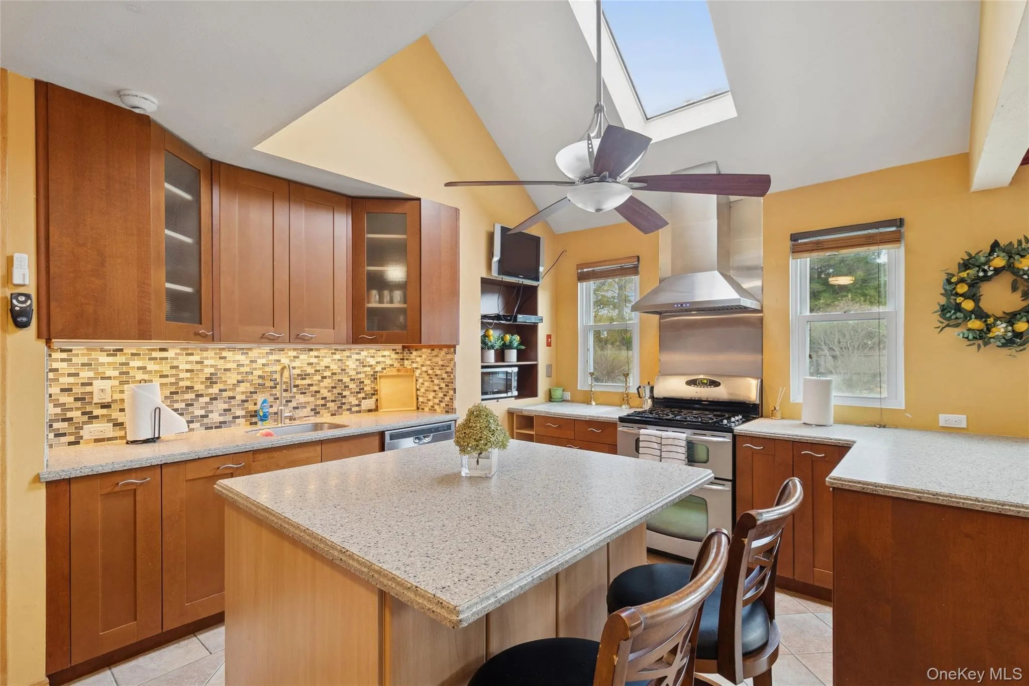 Kitchen with tasteful backsplash, brown cabinets, light tile patterned floors, stainless steel appliances, and wall chimney range hood Kitchen with tasteful backsplash, brown cabinets, light tile patterned floors, stainless steel appliances, and wall chimney range hood