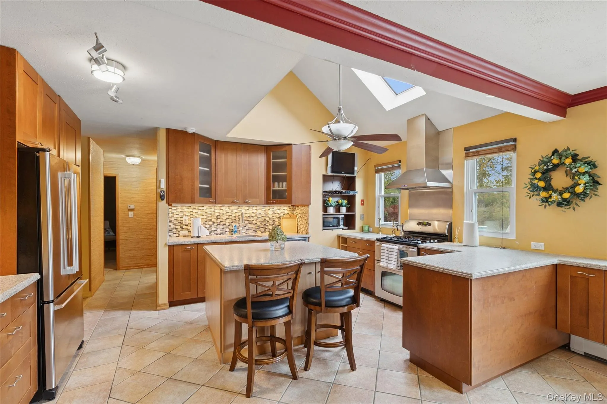 Kitchen featuring brown cabinetry, a skylight, decorative backsplash, island range hood, and stainless steel appliances Kitchen featuring brown cabinetry, a skylight, decorative backsplash, island range hood, and stainless steel appliances