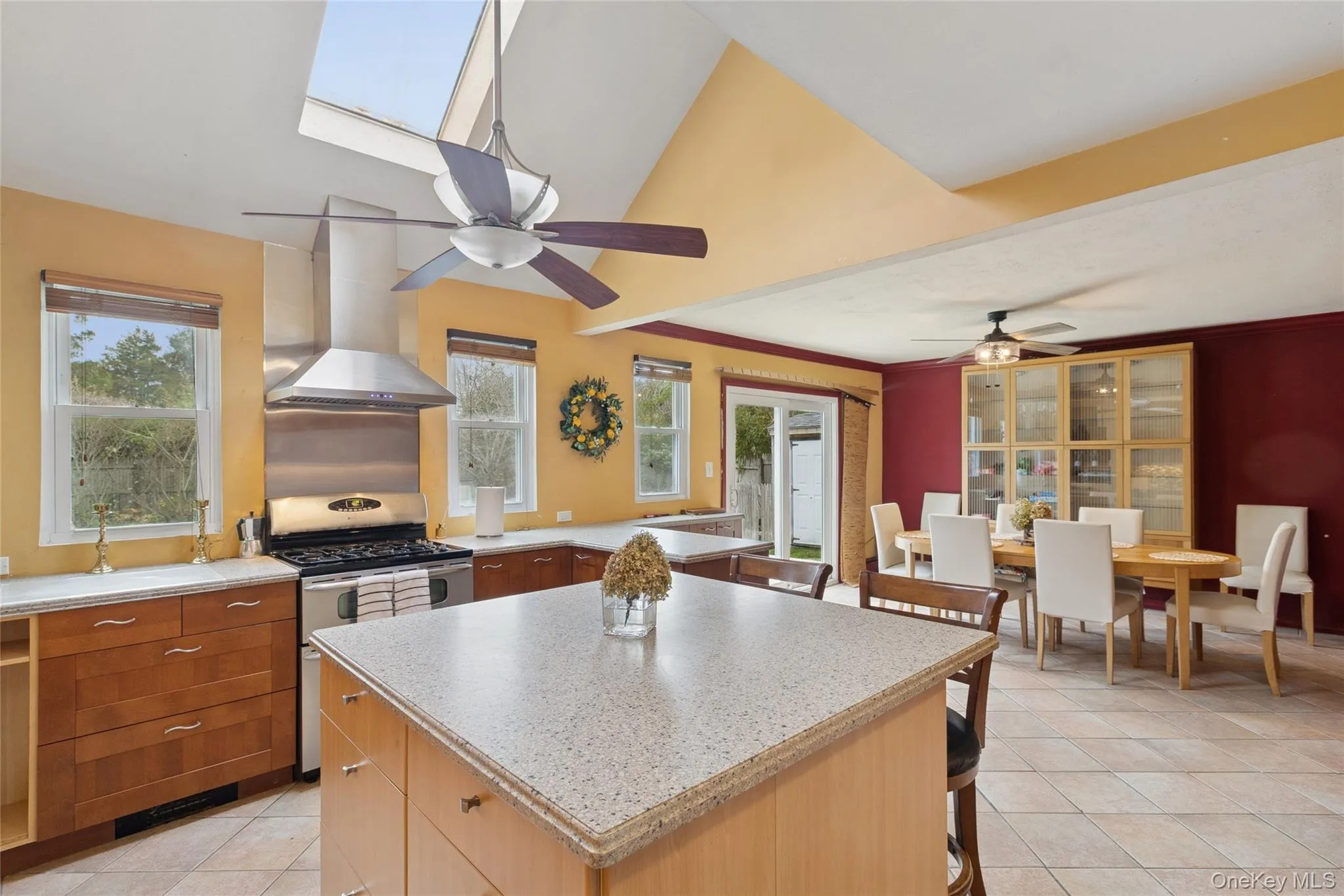 Kitchen with light tile patterned floors, wall chimney exhaust hood, a kitchen island, stainless steel range with gas cooktop, and a skylight Kitchen with light tile patterned floors, wall chimney exhaust hood, a kitchen island, stainless steel range with gas cooktop, and a skylight
