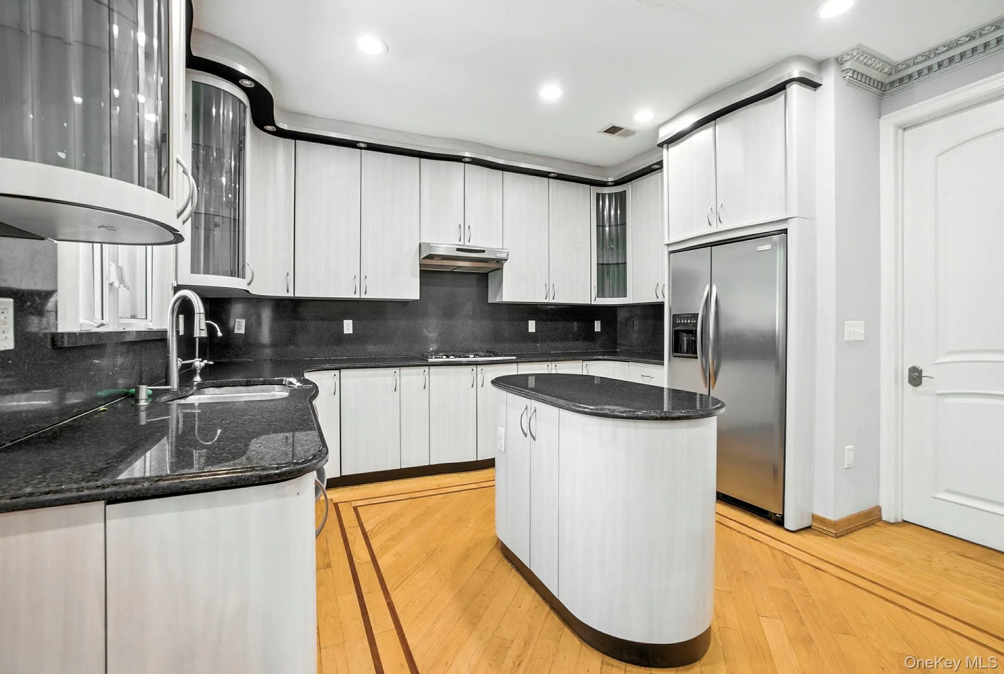 Kitchen with glass insert cabinets, backsplash, dark stone counters, stainless steel fridge, and white cabinetry Kitchen with glass insert cabinets, backsplash, dark stone counters, stainless steel fridge, and white cabinetry