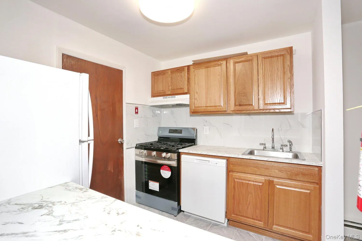 Kitchen featuring white appliances, decorative backsplash, under cabinet range hood, a baseboard heating unit, and brown cabinets Kitchen featuring white appliances, decorative backsplash, under cabinet range hood, a baseboard heating unit, and brown cabinets