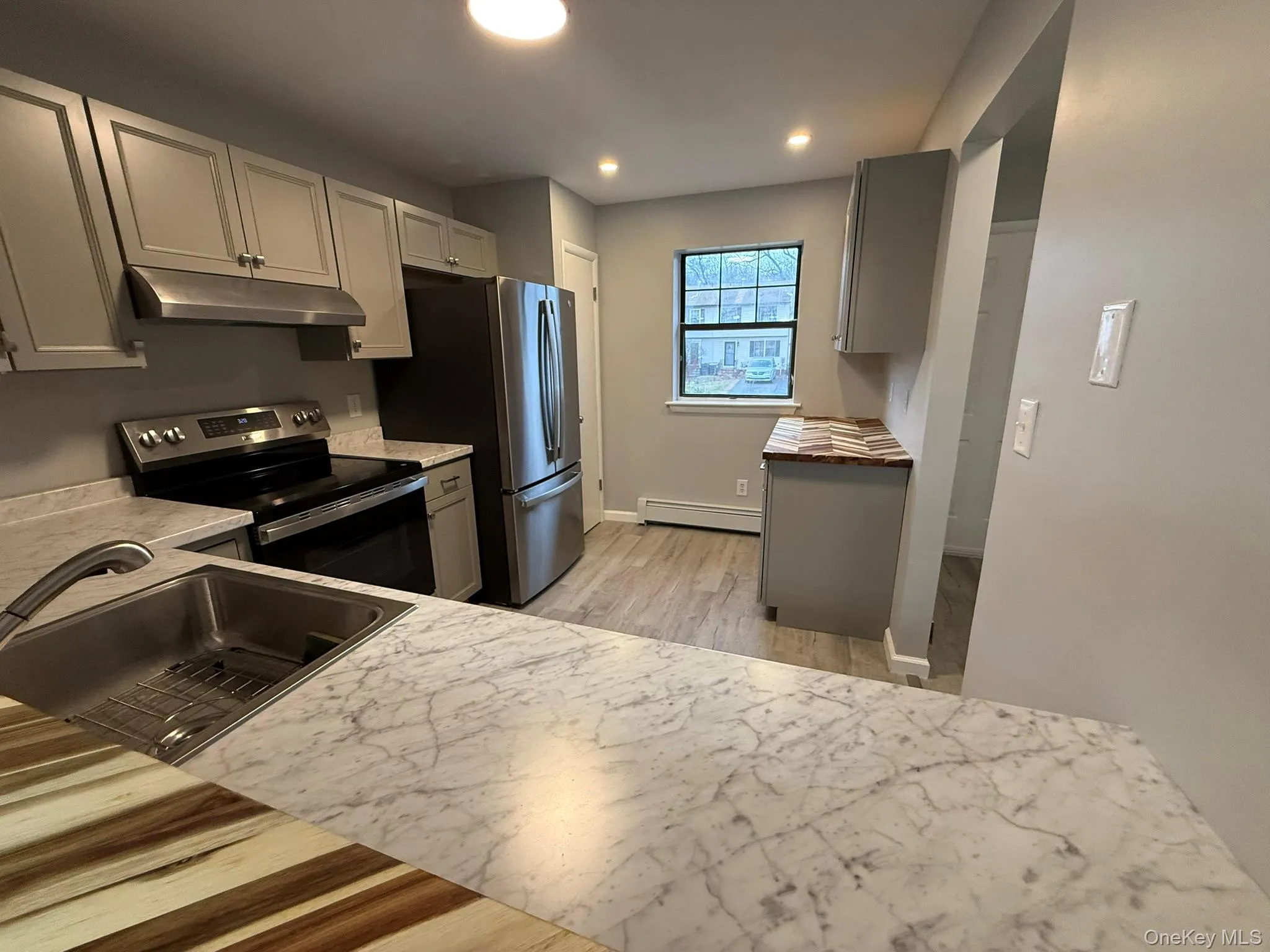 Kitchen featuring gray cabinetry, stainless steel electric stove, under cabinet range hood, a baseboard heating unit, and light wood-type flooring Kitchen featuring gray cabinetry, stainless steel electric stove, under cabinet range hood, a baseboard heating unit, and light wood-type flooring