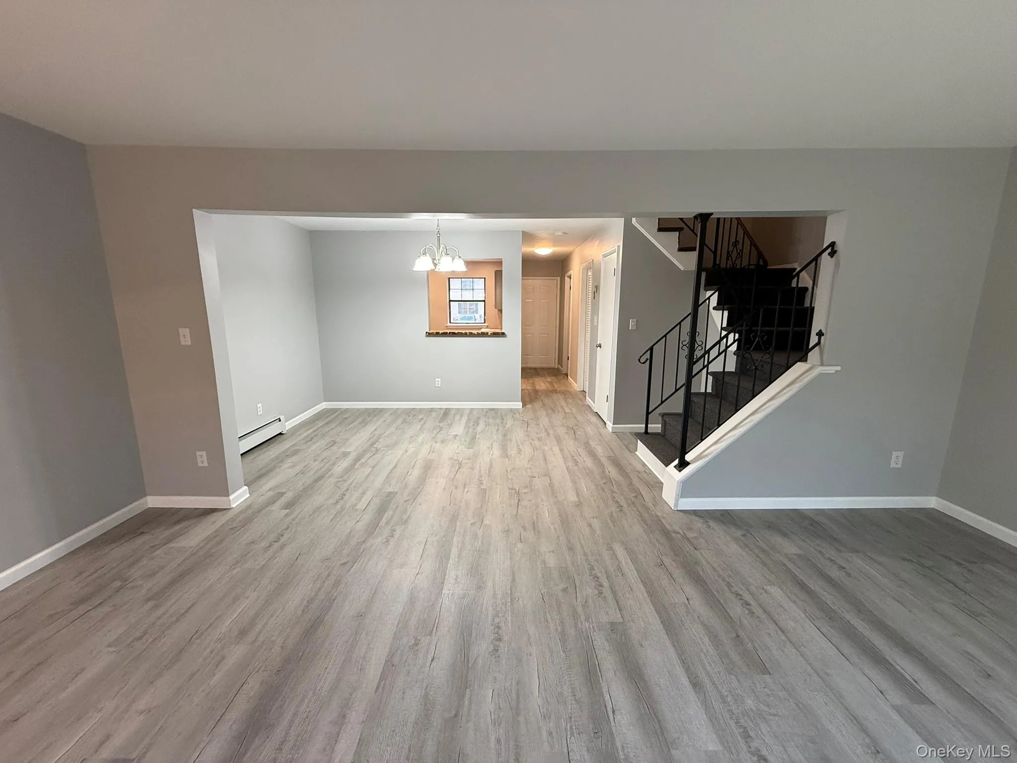 Unfurnished living room featuring a chandelier, light wood finished floors, stairway, and baseboard heating Unfurnished living room featuring a chandelier, light wood finished floors, stairway, and baseboard heating