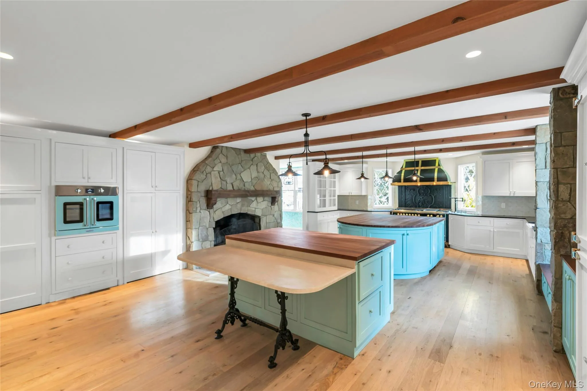 Expansive kitchen featuring exposed wood beams, light wood-finish flooring, a stone fireplace, white cabinetry, and two cherry wood kitchen islands Expansive kitchen featuring exposed wood beams, light wood-finish flooring, a stone fireplace, white cabinetry, and two cherry wood kitchen islands