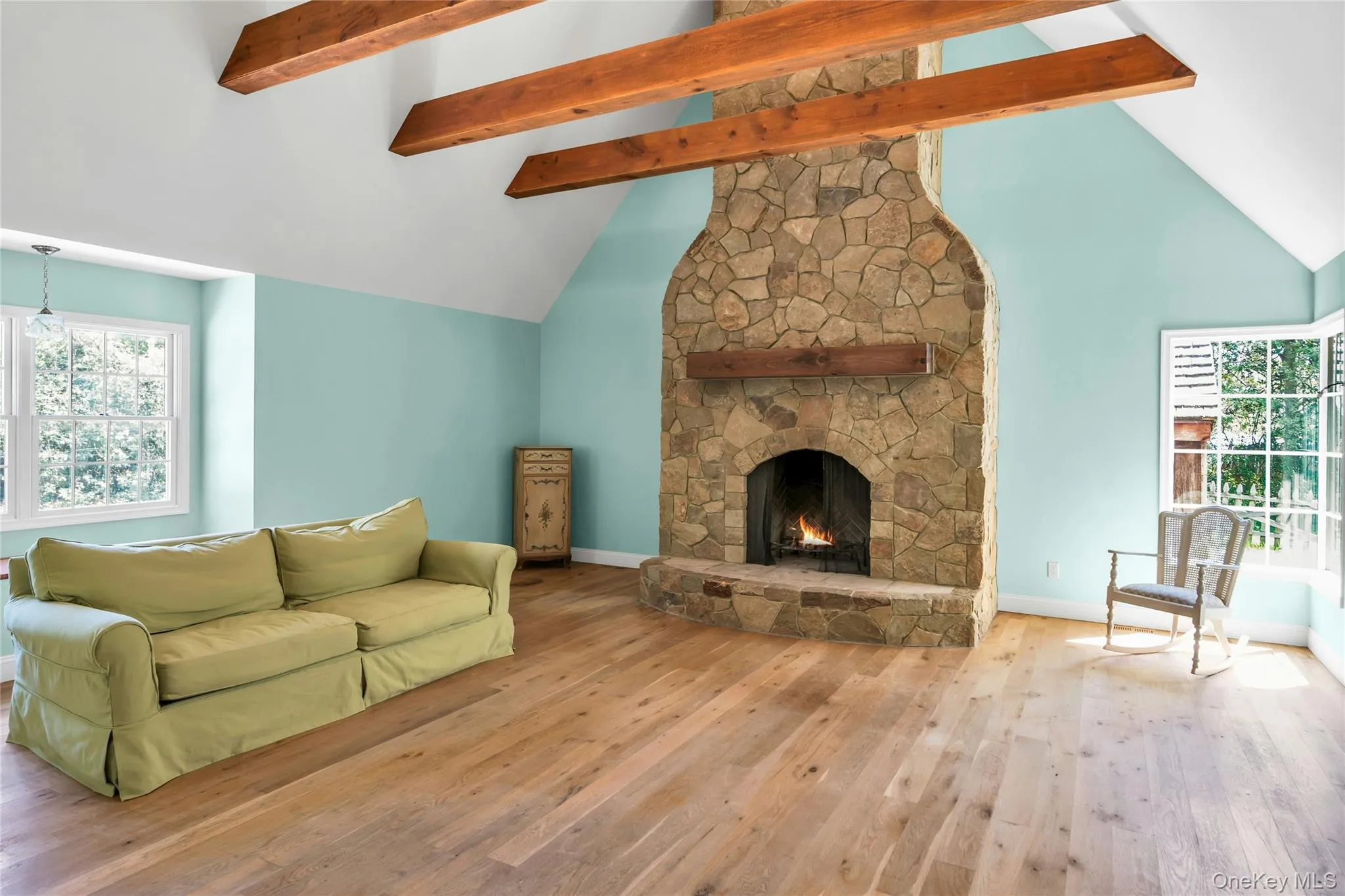 Spacious living area featuring a stone fireplace with a wood mantle, vaulted ceiling with exposed wood beams, wood-finish flooring, and multiple windows Spacious living area featuring a stone fireplace with a wood mantle, vaulted ceiling with exposed wood beams, wood-finish flooring, and multiple windows