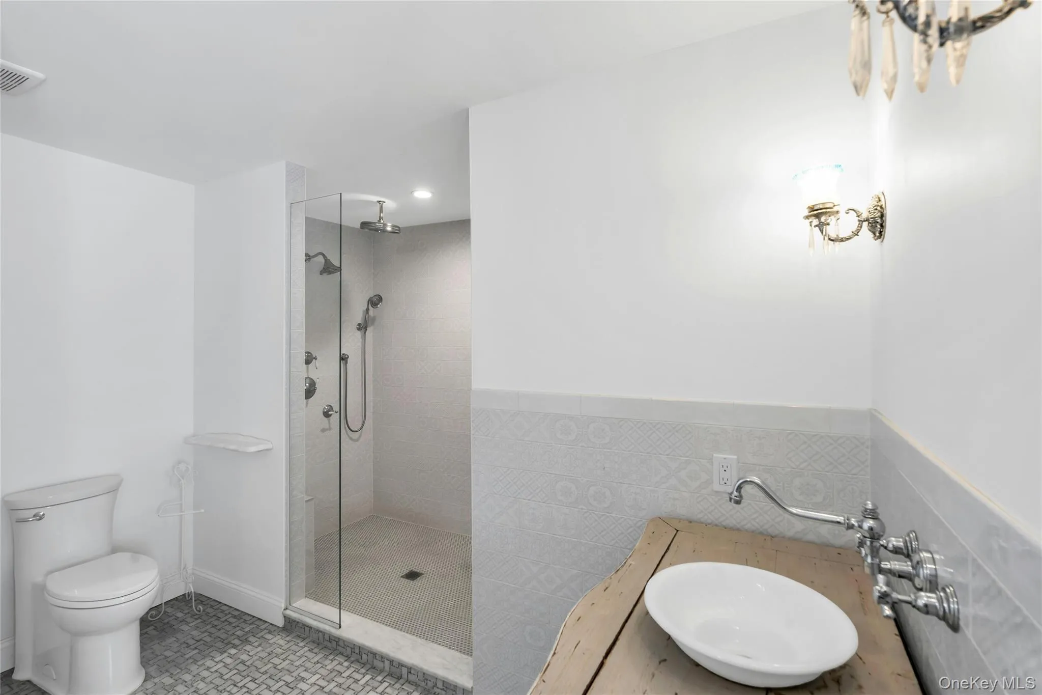 Bathroom featuring a glass-enclosed shower with multiple shower heads, a wall-mounted vanity with a vessel sink, decorative wall tile, and patterned floor tile Bathroom featuring a glass-enclosed shower with multiple shower heads, a wall-mounted vanity with a vessel sink, decorative wall tile, and patterned floor tile
