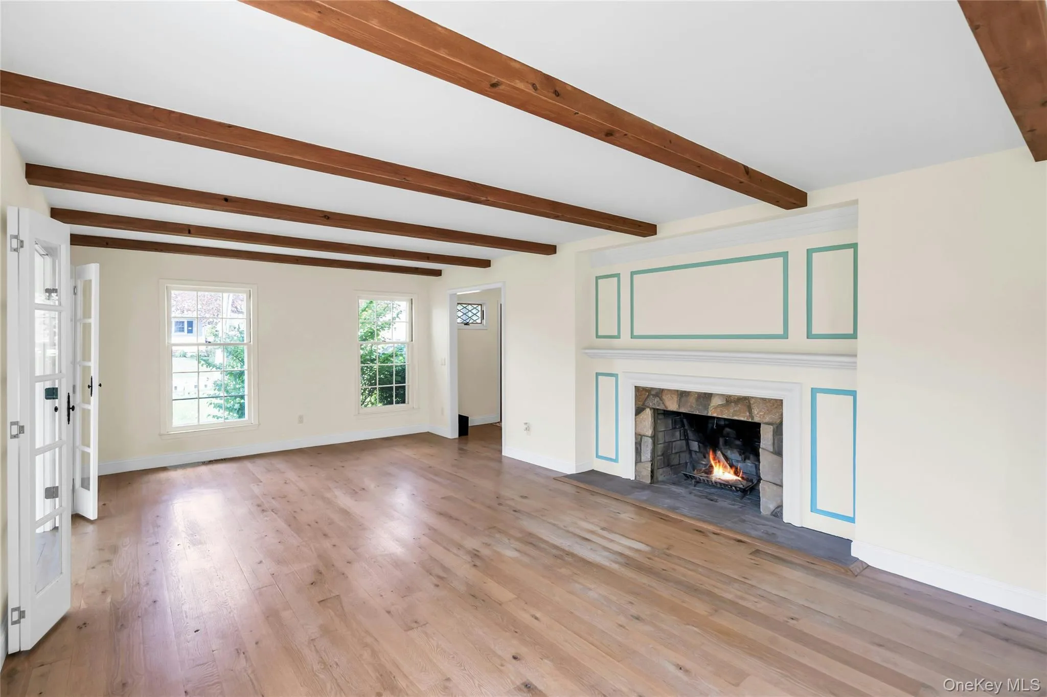 Living area featuring a stone fireplace with a decorative mantel, exposed wood ceiling beams, wood-finish flooring, and multiple windows Living area featuring a stone fireplace with a decorative mantel, exposed wood ceiling beams, wood-finish flooring, and multiple windows