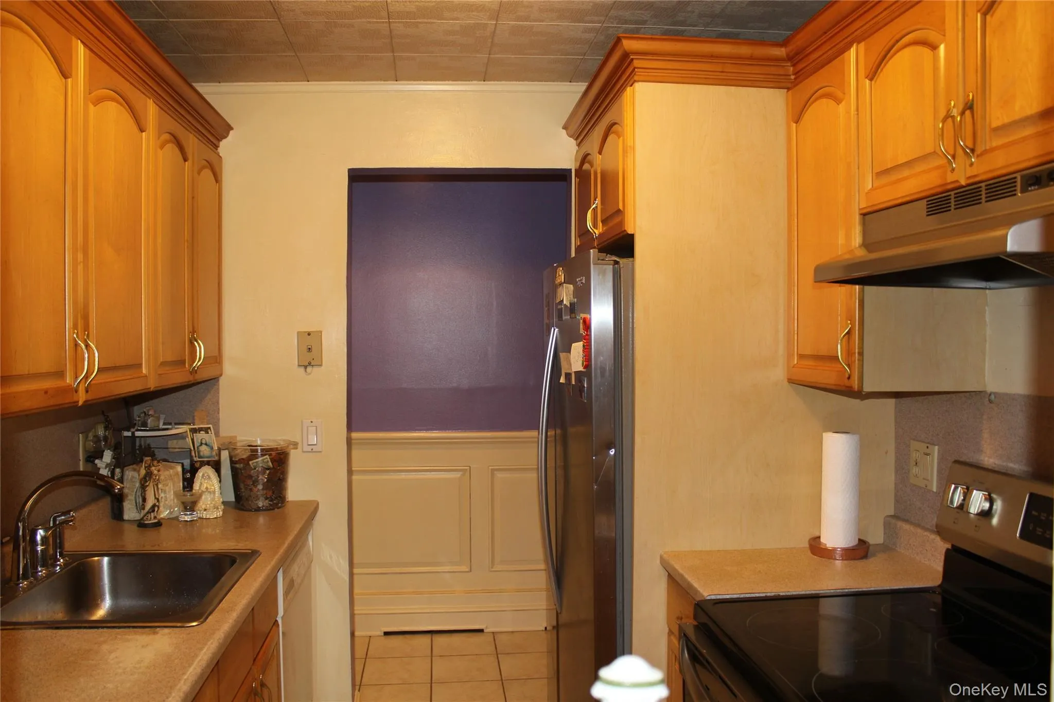 Kitchen with appliances with stainless steel finishes, light tile patterned floors, under cabinet range hood, light countertops, and brown cabinetry Kitchen with appliances with stainless steel finishes, light tile patterned floors, under cabinet range hood, light countertops, and brown cabinetry