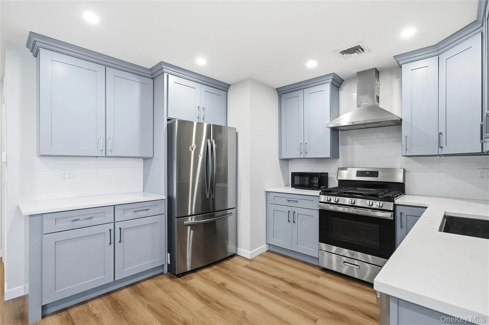 Kitchen with stainless steel appliances, wall chimney range hood, tasteful backsplash, gray cabinets, and light wood-style flooring Kitchen with stainless steel appliances, wall chimney range hood, tasteful backsplash, gray cabinets, and light wood-style flooring