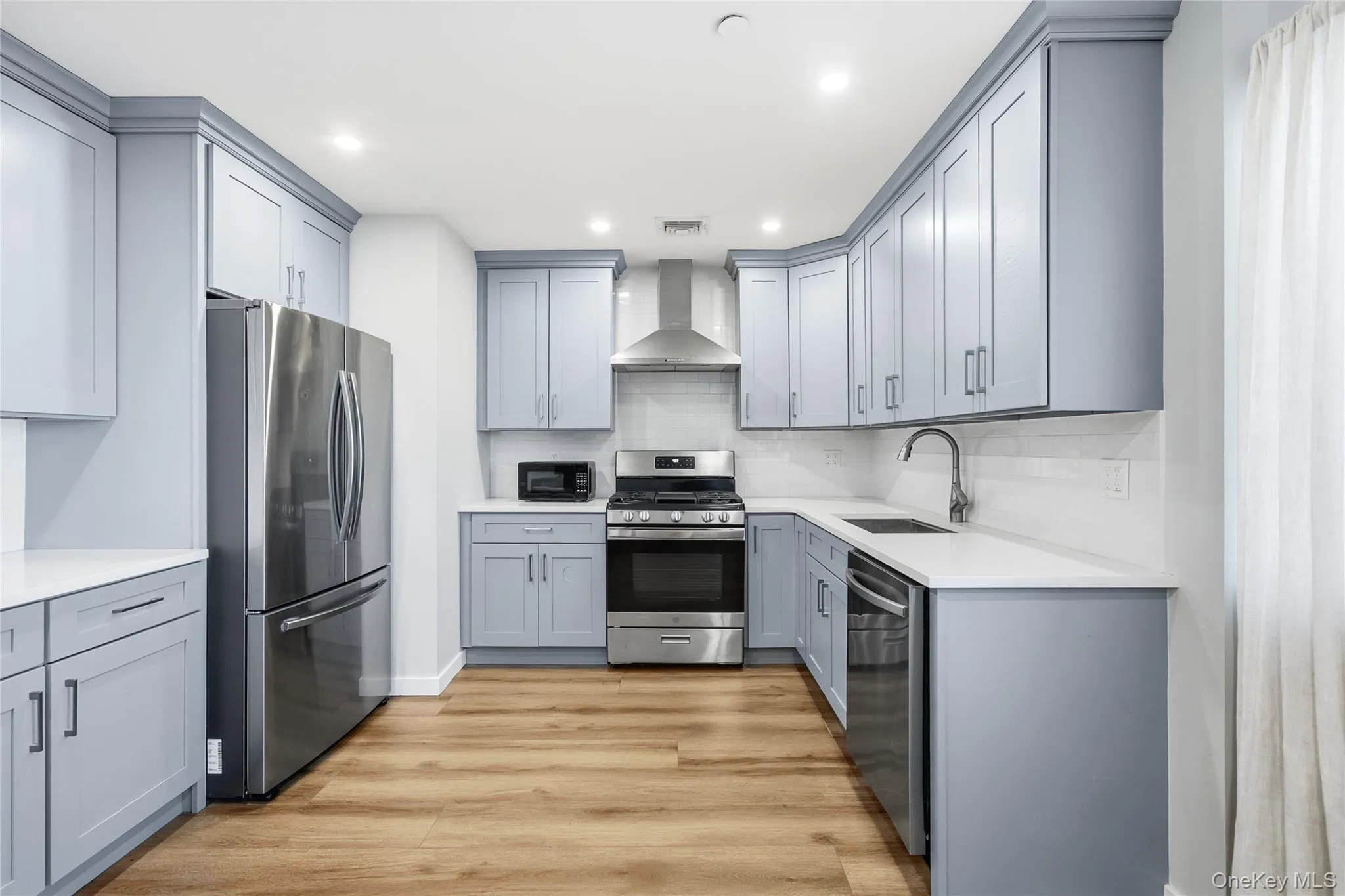 Kitchen with gray cabinets, appliances with stainless steel finishes, light wood-type flooring, light stone countertops, and wall chimney range hood Kitchen with gray cabinets, appliances with stainless steel finishes, light wood-type flooring, light stone countertops, and wall chimney range hood