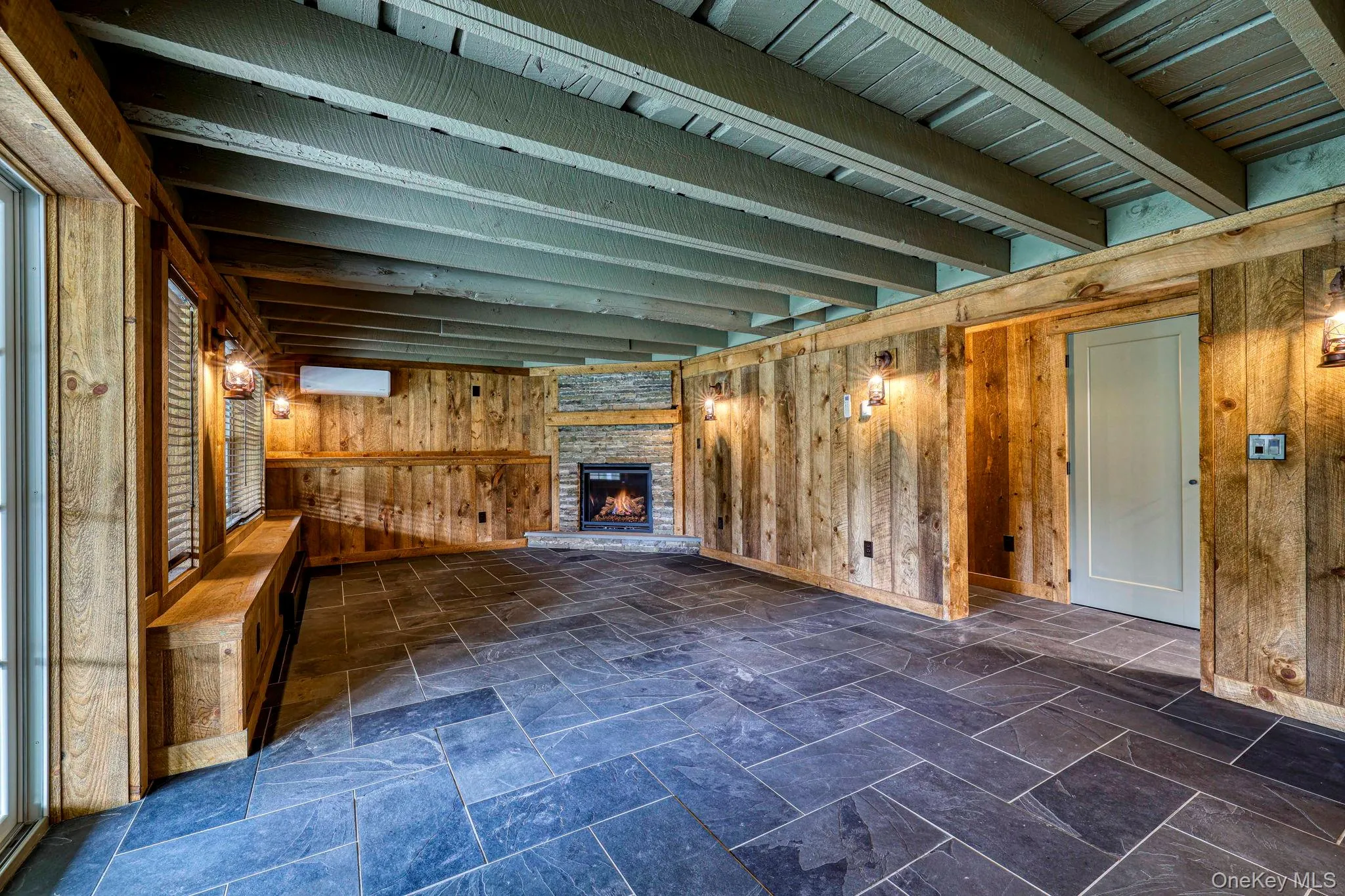 Unfurnished living room with wooden walls, beam ceiling, stone tile flooring, and a fireplace Unfurnished living room with wooden walls, beam ceiling, stone tile flooring, and a fireplace