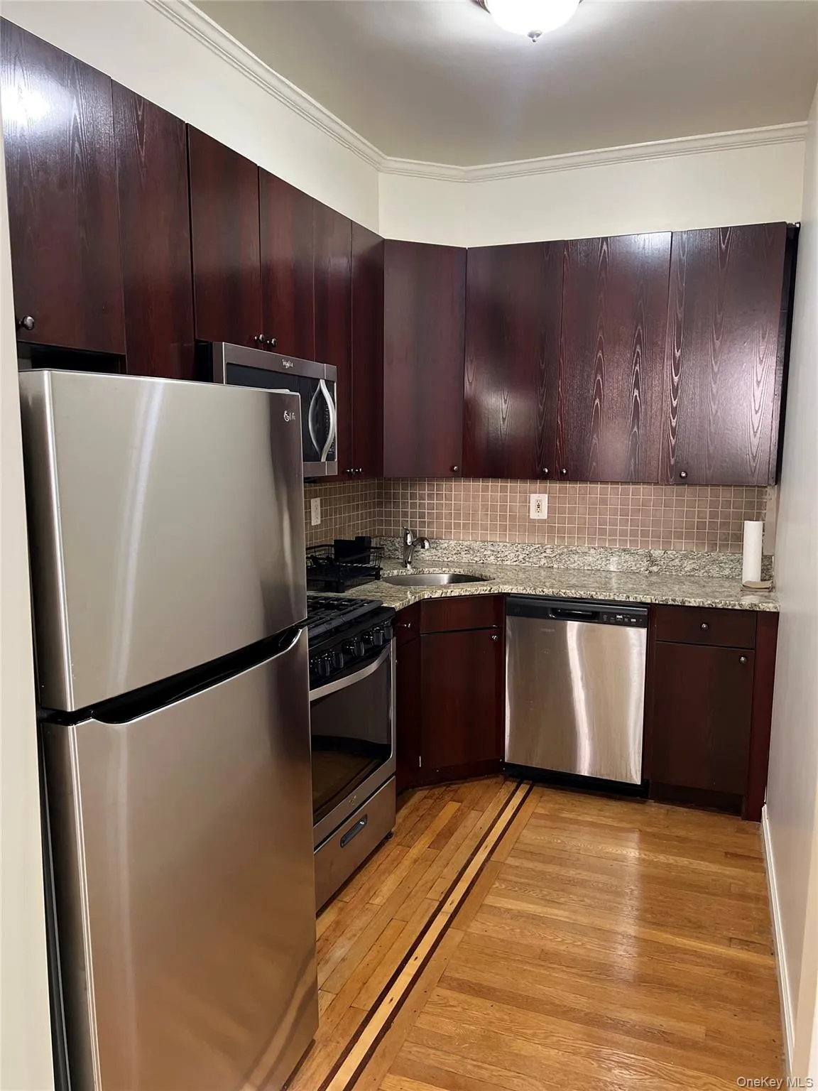 Kitchen with appliances with stainless steel finishes, dark brown cabinets, light wood-type flooring, tasteful backsplash, and light stone counters Kitchen with appliances with stainless steel finishes, dark brown cabinets, light wood-type flooring, tasteful backsplash, and light stone counters