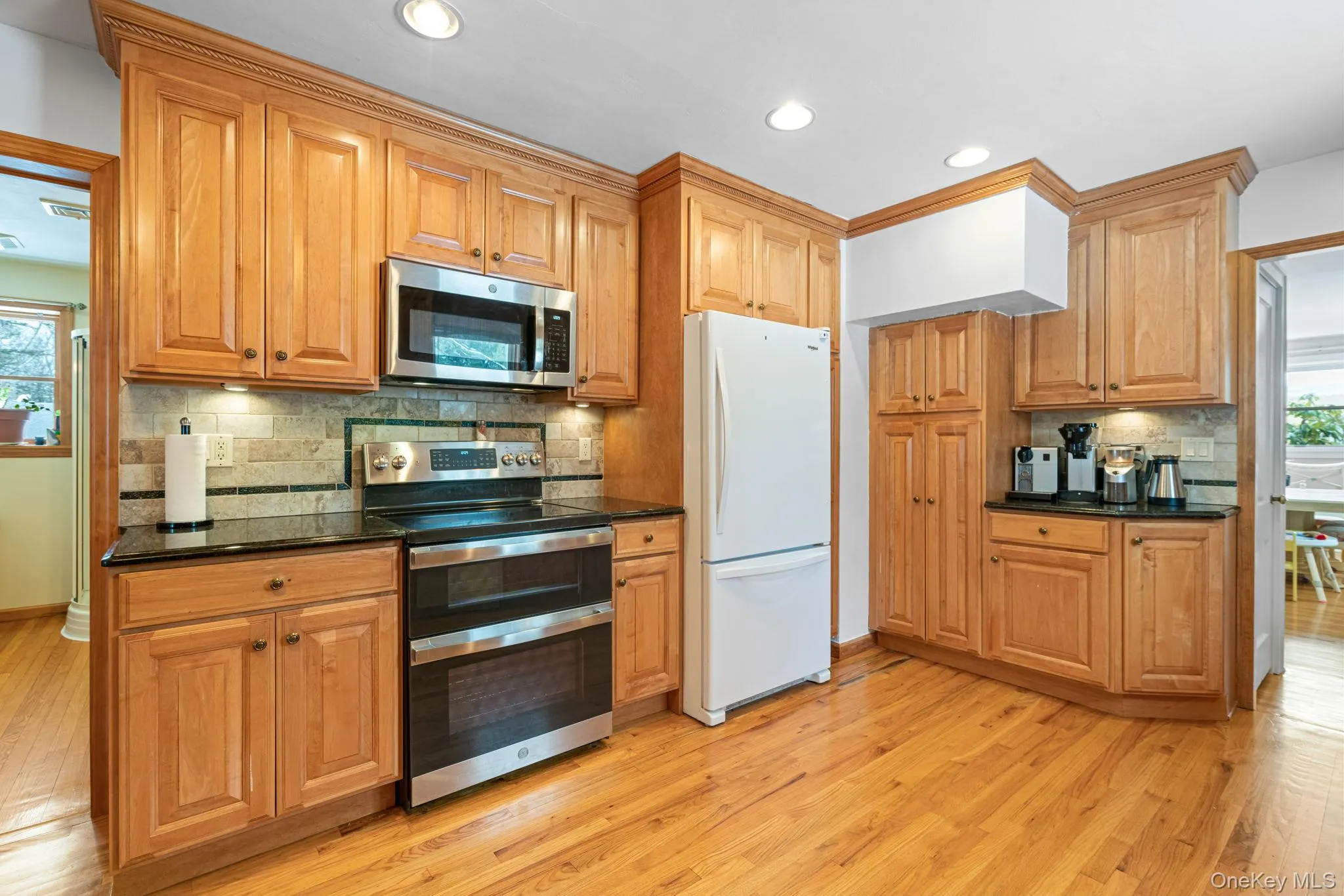 Kitchen featuring stainless steel appliances, backsplash, light wood finished floors, dark stone counters, and brown cabinetry Kitchen featuring stainless steel appliances, backsplash, light wood finished floors, dark stone counters, and brown cabinetry
