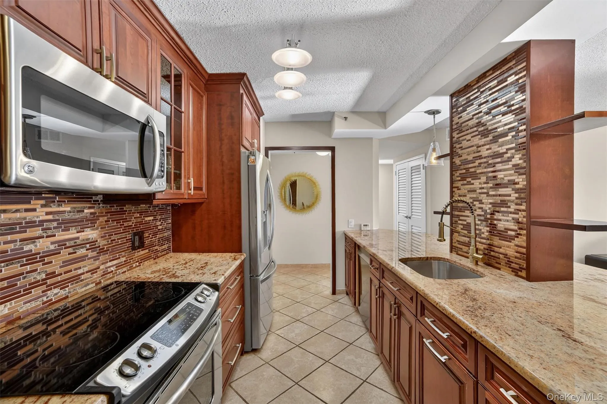 Kitchen with appliances with stainless steel finishes, glass insert cabinets, brown cabinets, light stone countertops, and a textured ceiling Kitchen with appliances with stainless steel finishes, glass insert cabinets, brown cabinets, light stone countertops, and a textured ceiling