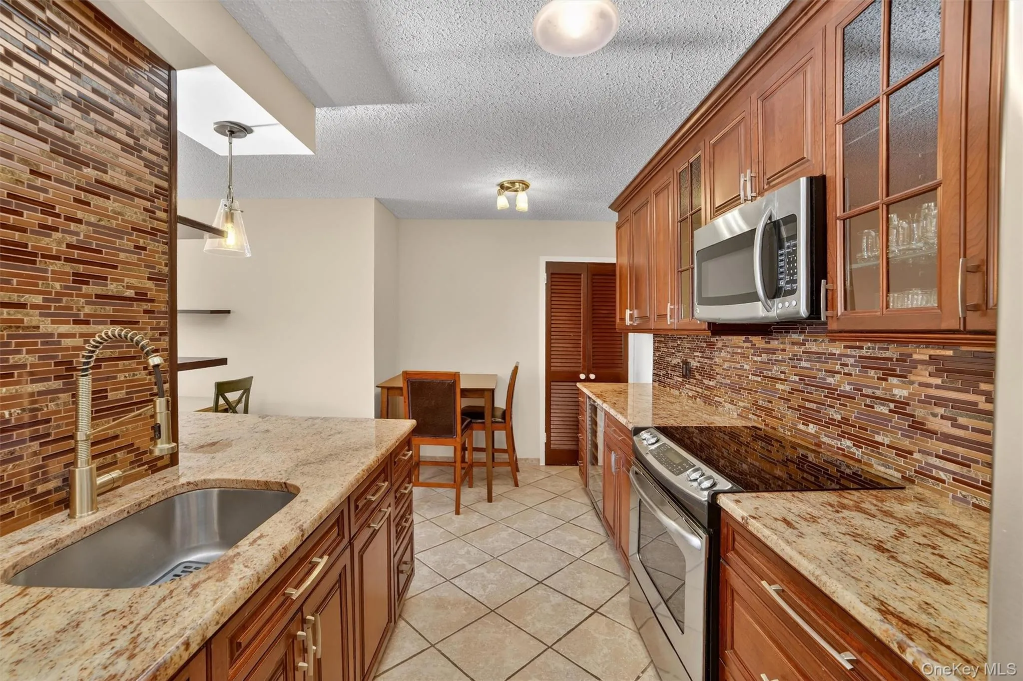 Kitchen featuring tasteful backsplash, appliances with stainless steel finishes, hanging light fixtures, brown cabinetry, and a textured ceiling Kitchen featuring tasteful backsplash, appliances with stainless steel finishes, hanging light fixtures, brown cabinetry, and a textured ceiling