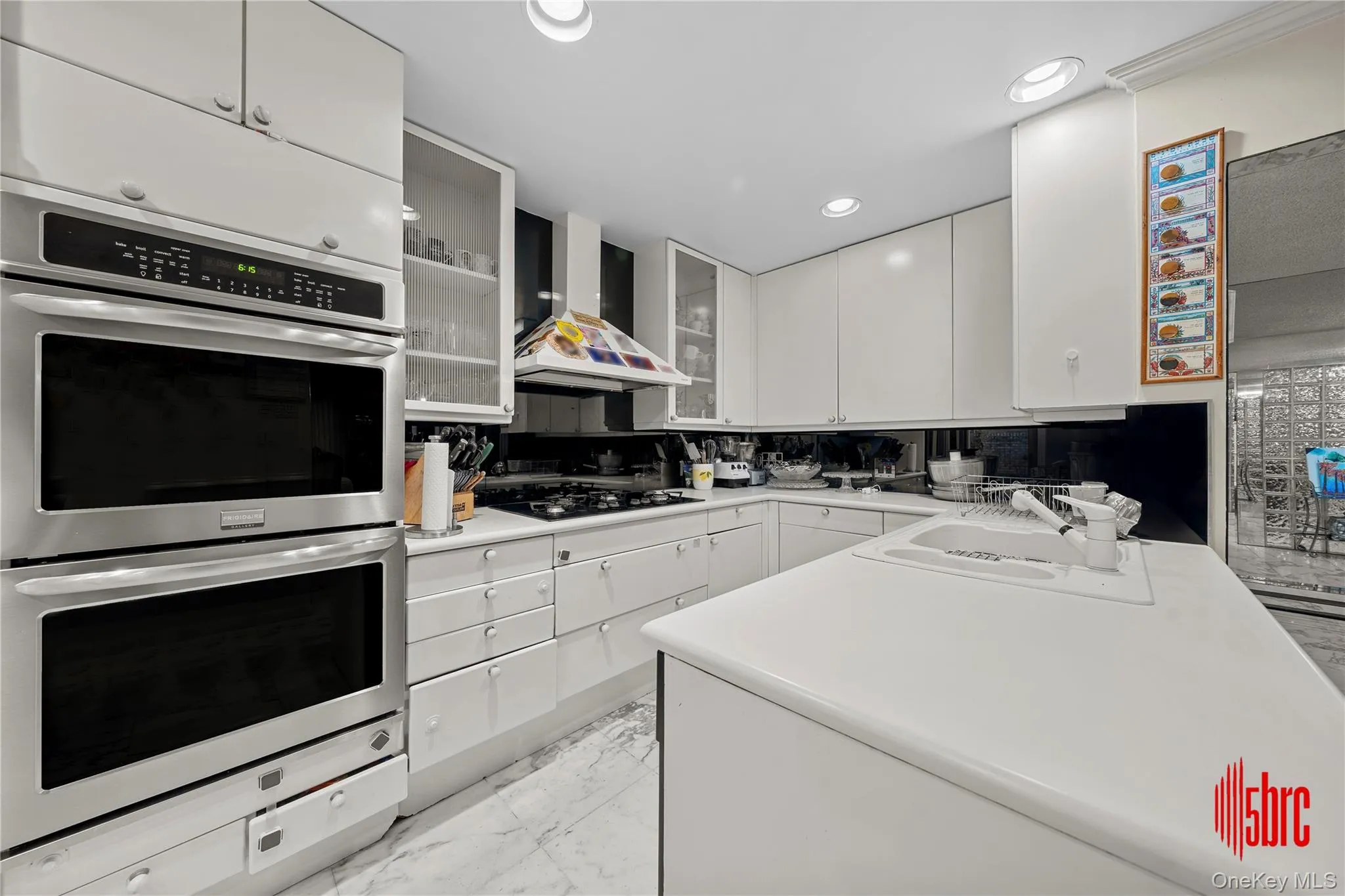 Kitchen with double oven, light countertops, white cabinets, wall chimney exhaust hood, and light marble finish flooring Kitchen with double oven, light countertops, white cabinets, wall chimney exhaust hood, and light marble finish flooring