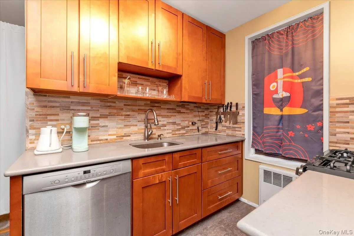 Kitchen with tasteful backsplash, dishwasher, brown cabinets, radiator heating unit, and light stone counters Kitchen with tasteful backsplash, dishwasher, brown cabinets, radiator heating unit, and light stone counters