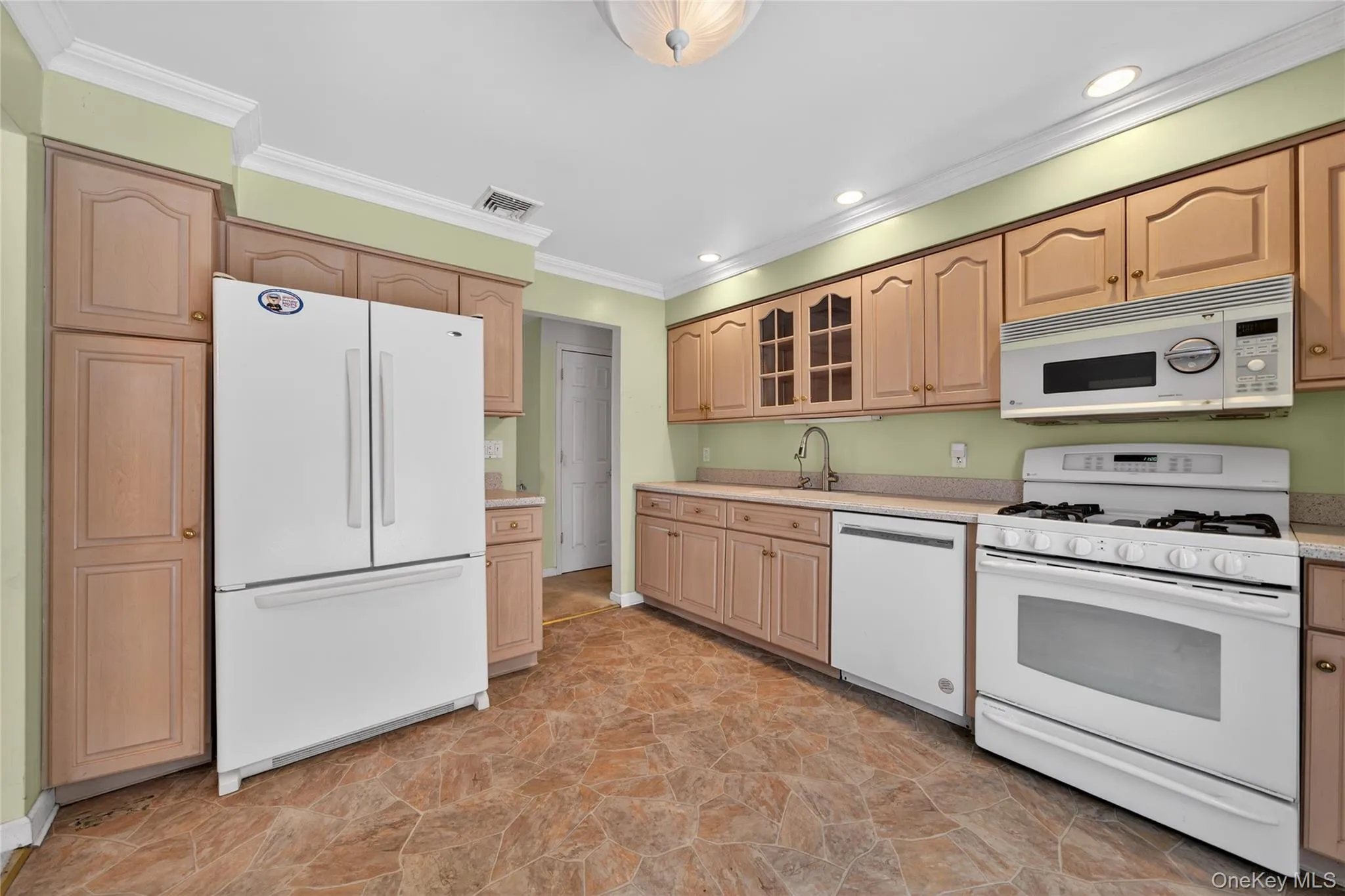 Kitchen with white appliances, stone finish flooring, glass insert cabinets, light brown cabinetry, and ornamental molding Kitchen with white appliances, stone finish flooring, glass insert cabinets, light brown cabinetry, and ornamental molding