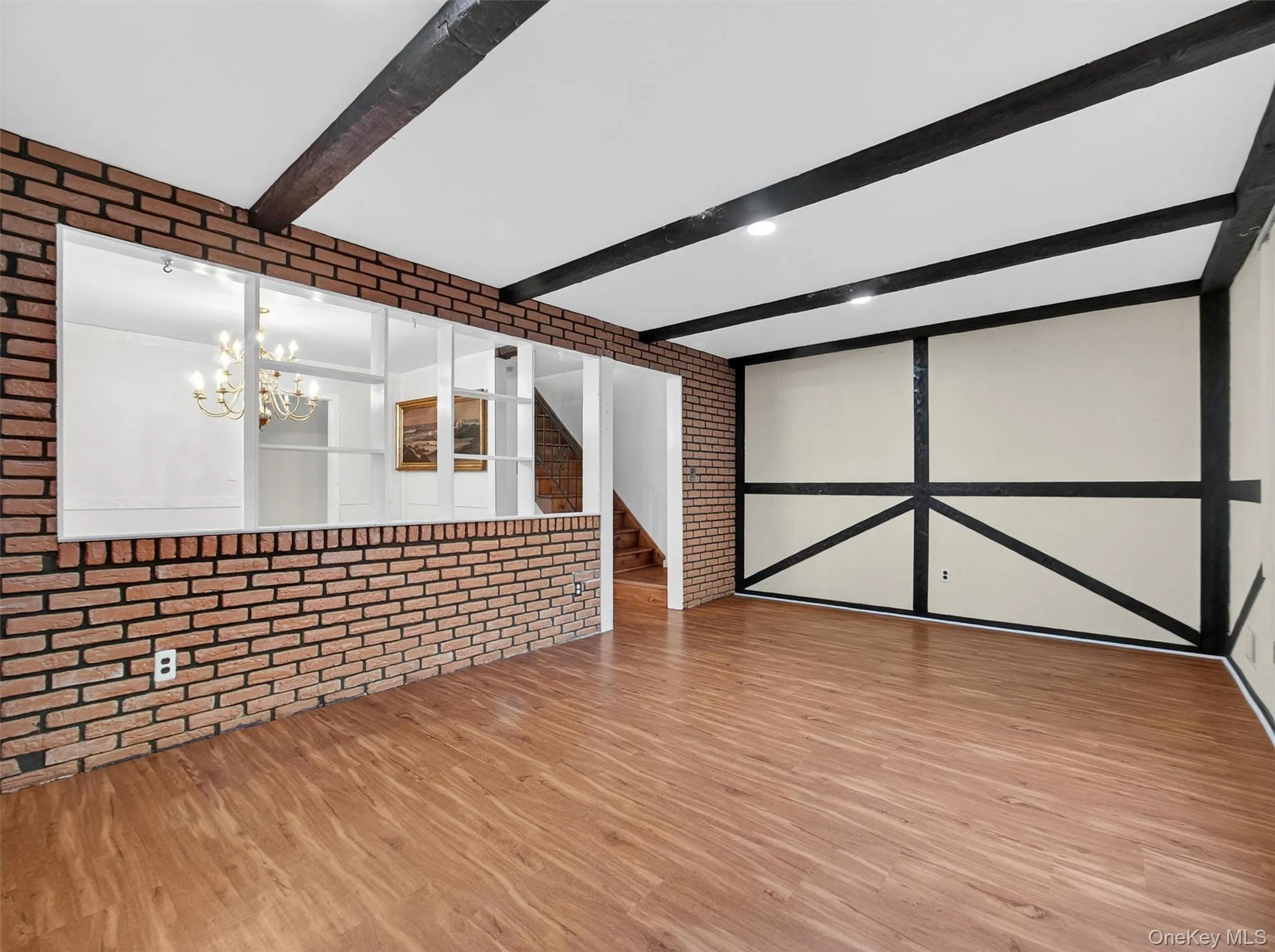 Unfurnished living room with brick wall, stairs, wood finished floors, a chandelier, and beam ceiling Unfurnished living room with brick wall, stairs, wood finished floors, a chandelier, and beam ceiling