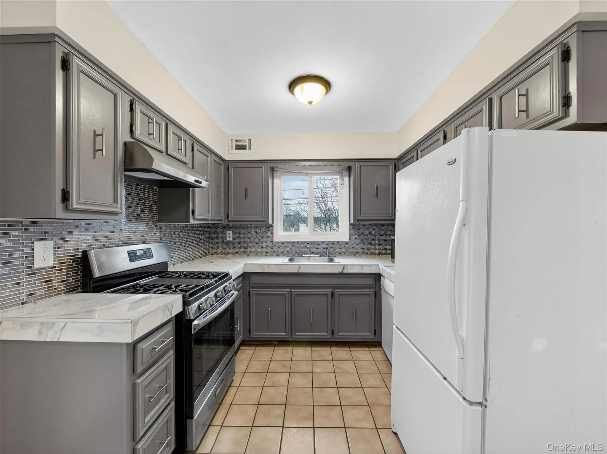 Kitchen featuring gray cabinets, freestanding refrigerator, stainless steel gas stove, under cabinet range hood, and tasteful backsplash Kitchen featuring gray cabinets, freestanding refrigerator, stainless steel gas stove, under cabinet range hood, and tasteful backsplash