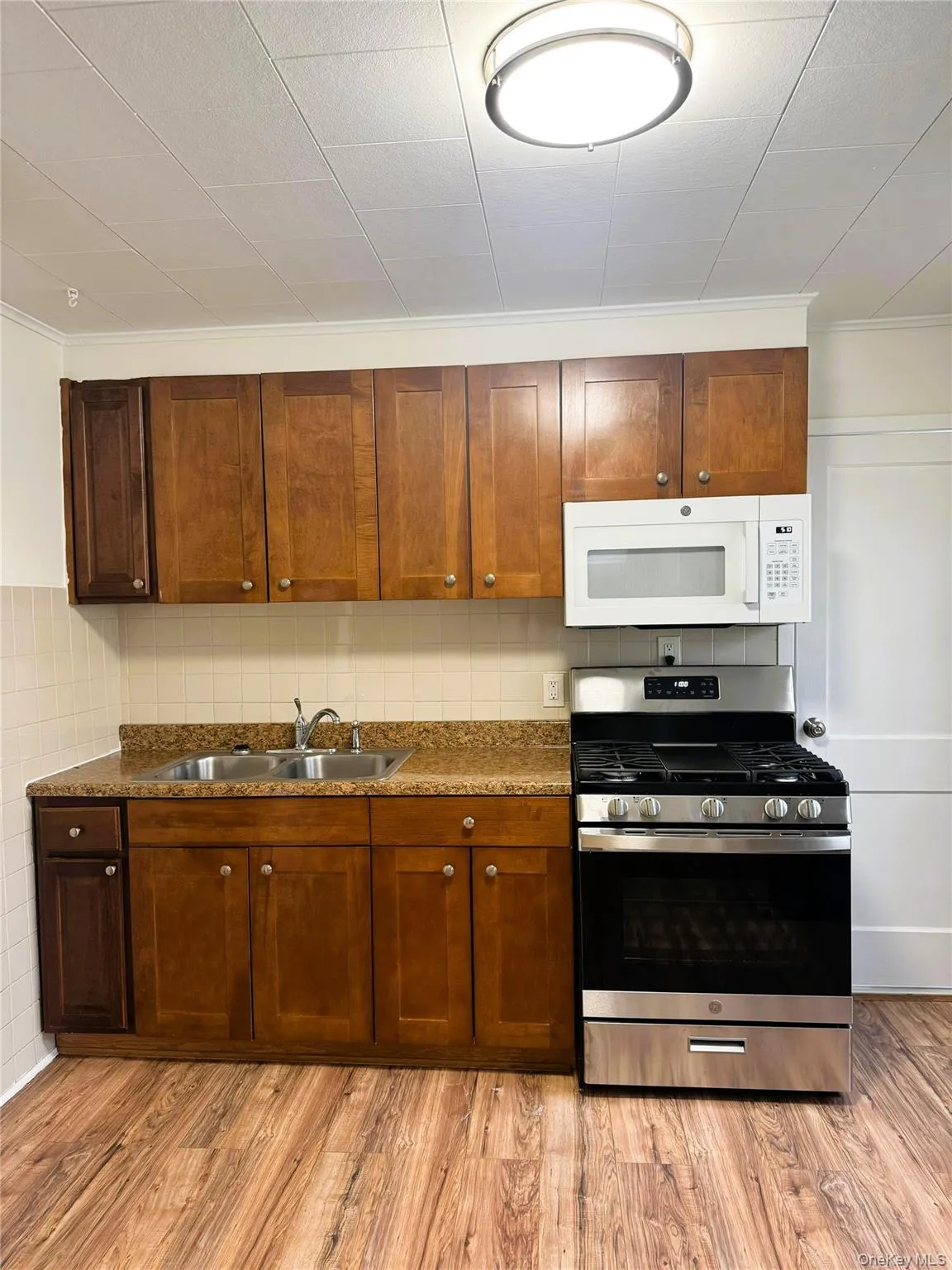 Kitchen with gas stove, white microwave, light wood finished floors, ornamental molding, and brown cabinetry Kitchen with gas stove, white microwave, light wood finished floors, ornamental molding, and brown cabinetry