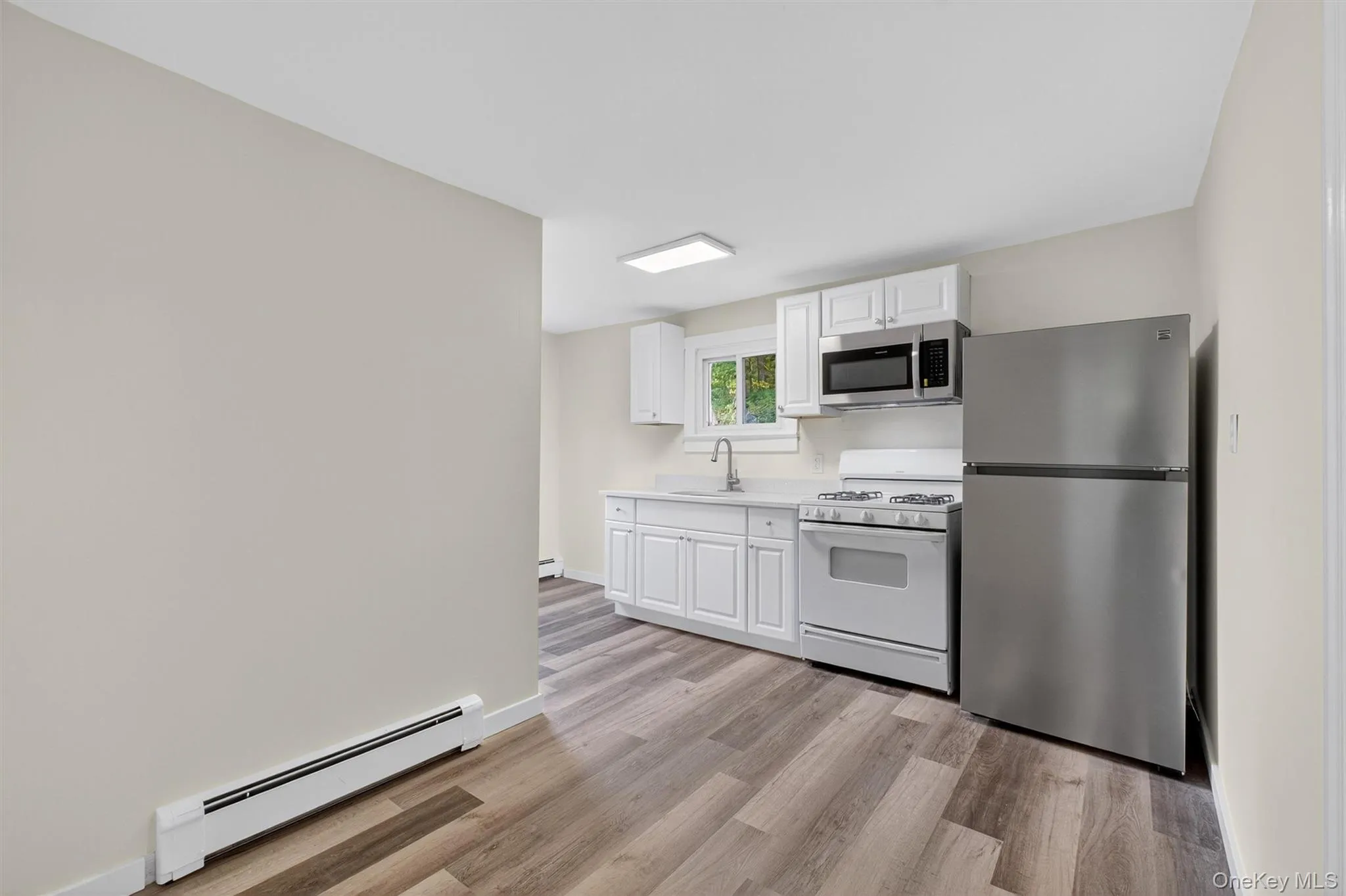 Kitchen featuring a baseboard heating unit, stainless steel appliances, light countertops, white cabinetry, and light wood-type flooring Kitchen featuring a baseboard heating unit, stainless steel appliances, light countertops, white cabinetry, and light wood-type flooring
