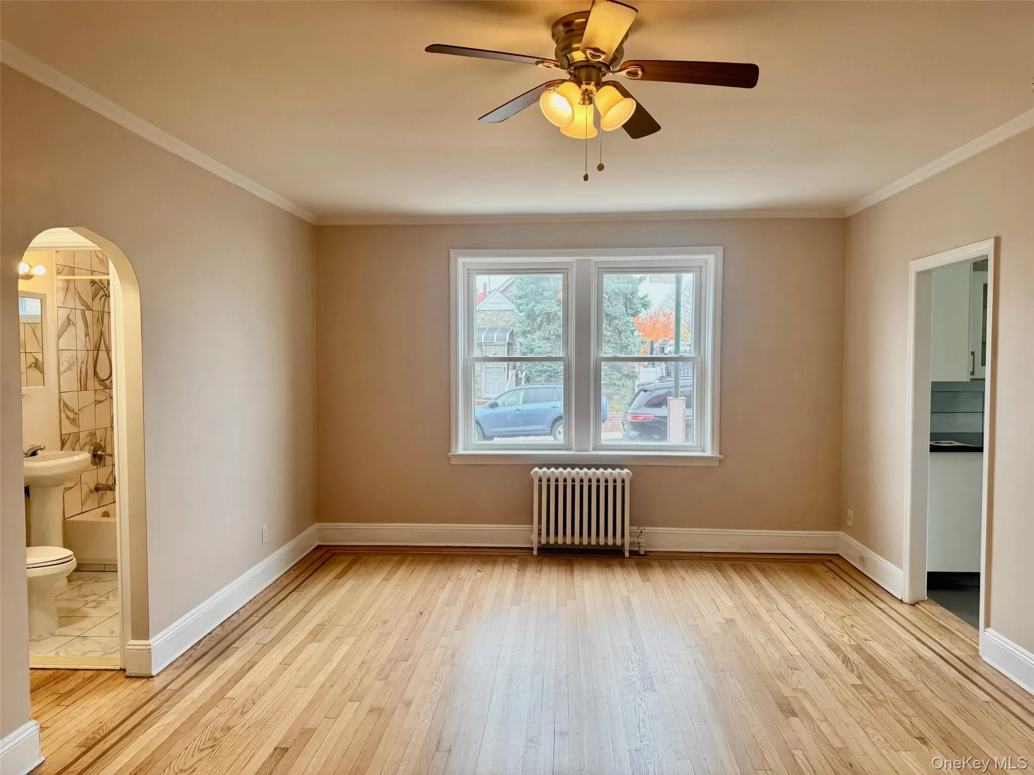 Empty room with radiator, light wood-style floors, arched walkways, crown molding, and a ceiling fan Empty room with radiator, light wood-style floors, arched walkways, crown molding, and a ceiling fan