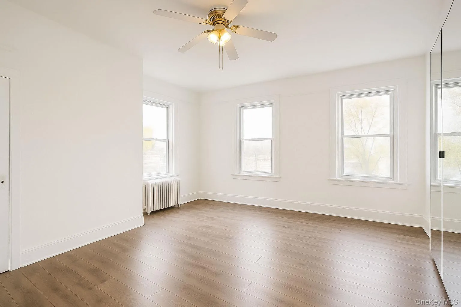 Empty room with radiator, light wood-style flooring, healthy amount of natural light, and a ceiling fan Empty room with radiator, light wood-style flooring, healthy amount of natural light, and a ceiling fan
