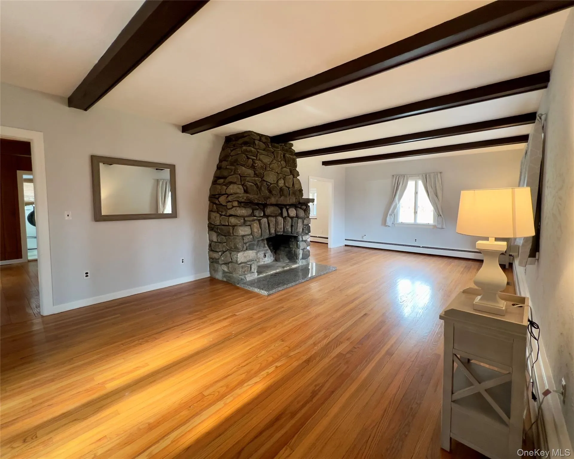 Unfurnished living room featuring hardwood flooring, beamed ceiling, a stone fireplace, and baseboard heating Unfurnished living room featuring hardwood flooring, beamed ceiling, a stone fireplace, and baseboard heating