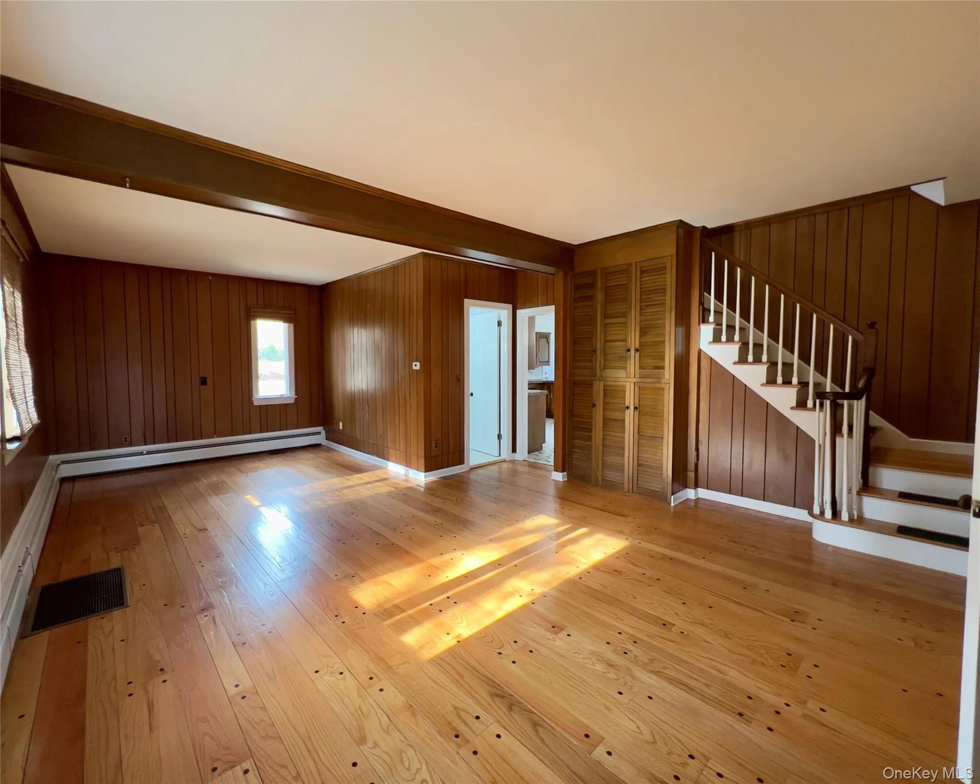 Unfurnished living room with hardwood flooring, wood walls, stairs, beam ceiling, and baseboard heating Unfurnished living room with hardwood flooring, wood walls, stairs, beam ceiling, and baseboard heating