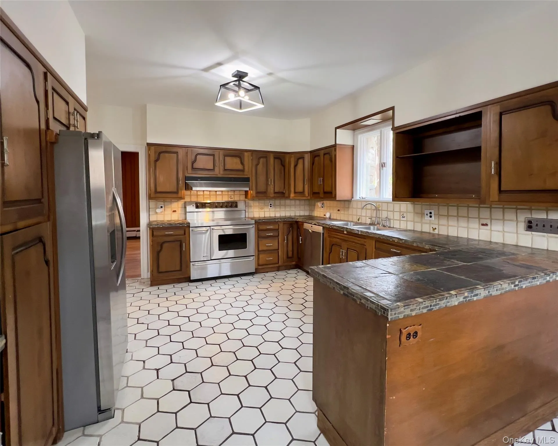 Kitchen with open shelves, stainless steel appliances, a peninsula, decorative backsplash, stone tile countertops, brown cabinetry and baseboard heating Kitchen with open shelves, stainless steel appliances, a peninsula, decorative backsplash, stone tile countertops, brown cabinetry and baseboard heating