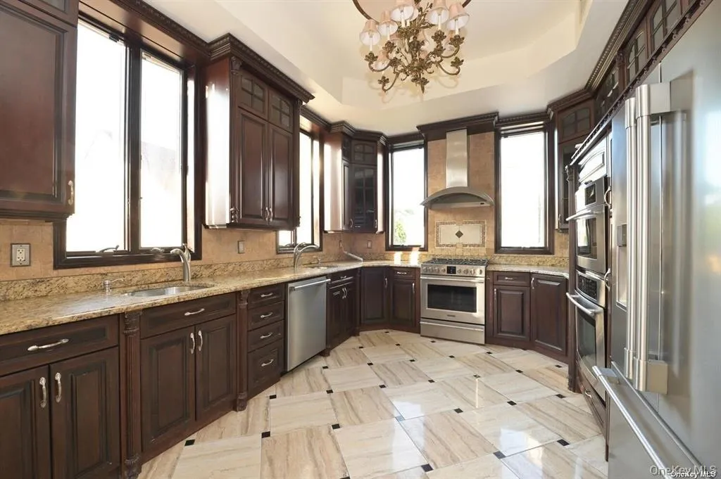 Kitchen with dark brown cabinetry, a raised ceiling, high end appliances, and wall chimney exhaust hood Kitchen with dark brown cabinetry, a raised ceiling, high end appliances, and wall chimney exhaust hood