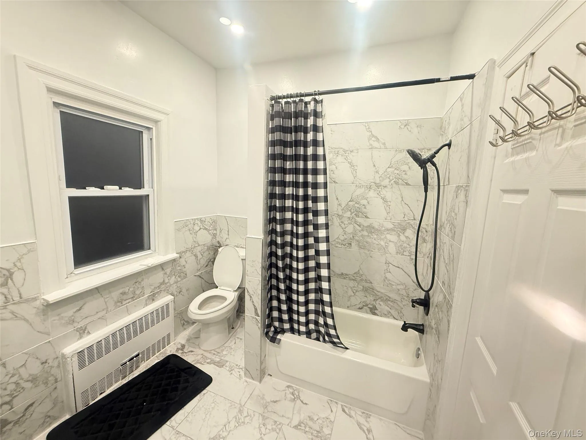 Full bathroom featuring shower / bathtub combination with curtain, radiator, tile walls, wainscoting, and light marble finish flooring Full bathroom featuring shower / bathtub combination with curtain, radiator, tile walls, wainscoting, and light marble finish flooring