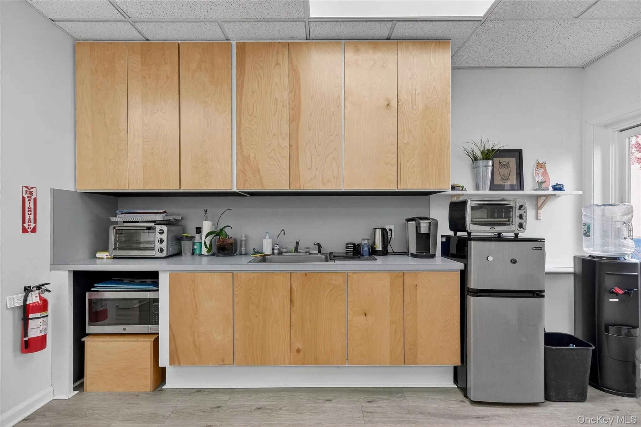 Kitchen featuring light brown cabinetry, light countertops, modern cabinets, appliances with stainless steel finishes, and a drop ceiling Kitchen featuring light brown cabinetry, light countertops, modern cabinets, appliances with stainless steel finishes, and a drop ceiling