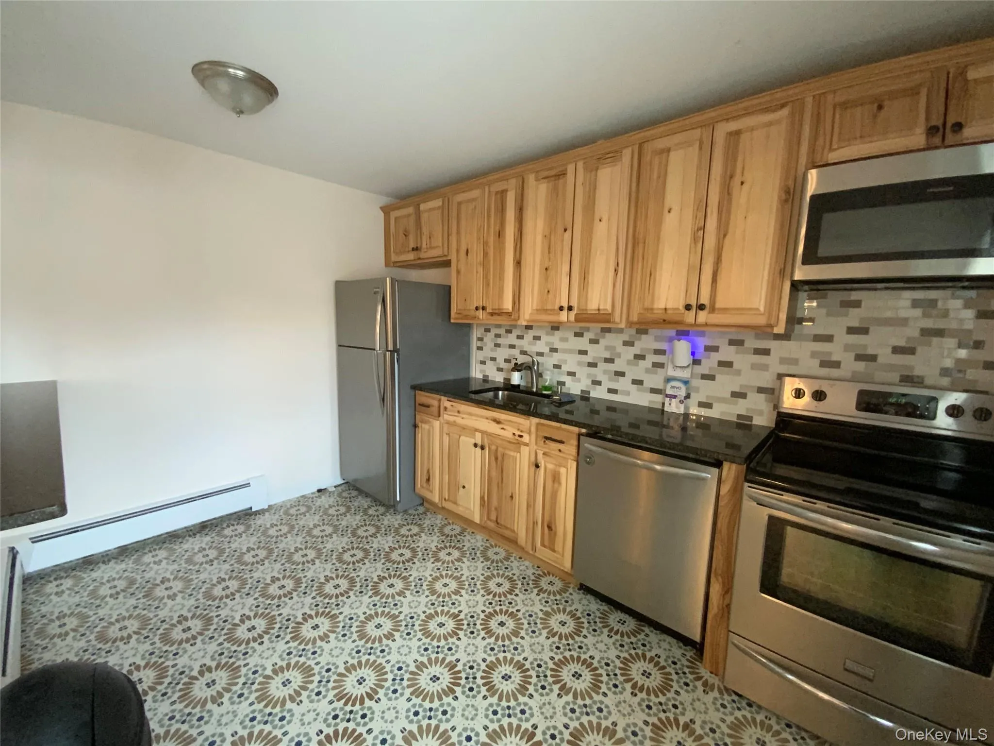 Kitchen with appliances with stainless steel finishes, backsplash, a baseboard radiator, dark stone countertops, and light flooring Kitchen with appliances with stainless steel finishes, backsplash, a baseboard radiator, dark stone countertops, and light flooring