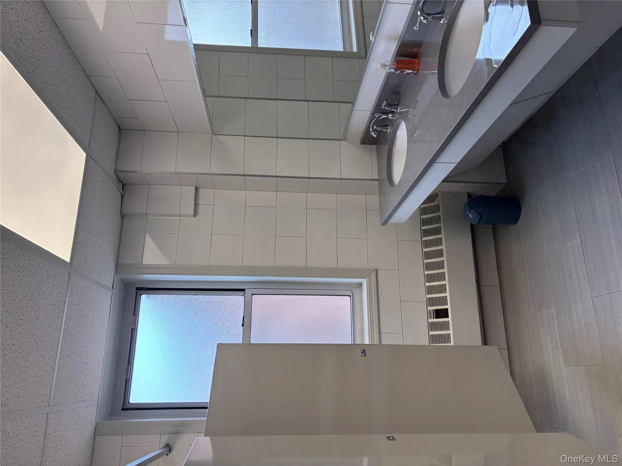 Full bathroom featuring tile walls, double vanity, a paneled ceiling, radiator, and light tile patterned floors Full bathroom featuring tile walls, double vanity, a paneled ceiling, radiator, and light tile patterned floors