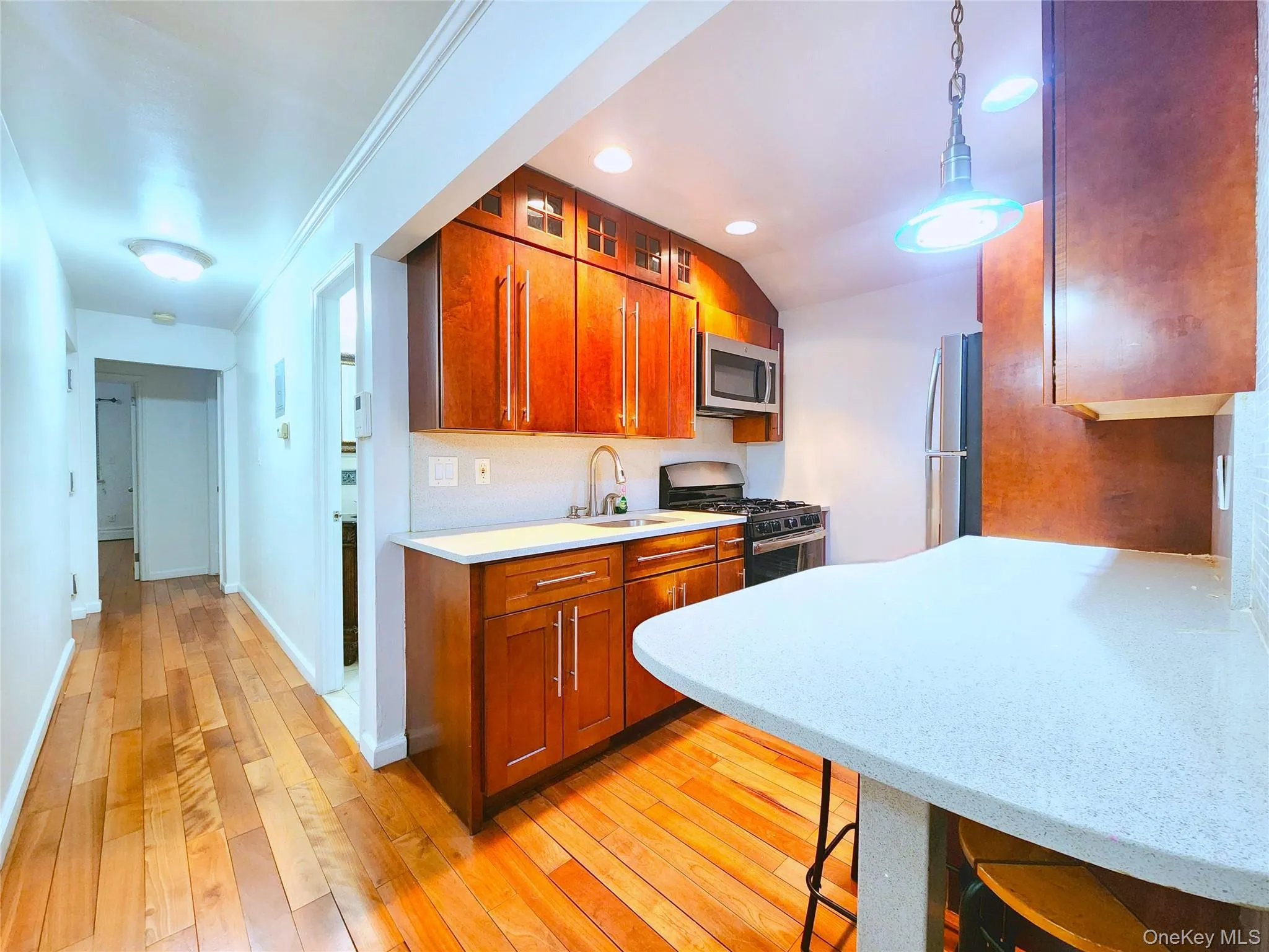 Kitchen with stainless steel appliances, hanging light fixtures, brown cabinetry, light wood-type flooring, and light stone countertops Kitchen with stainless steel appliances, hanging light fixtures, brown cabinetry, light wood-type flooring, and light stone countertops