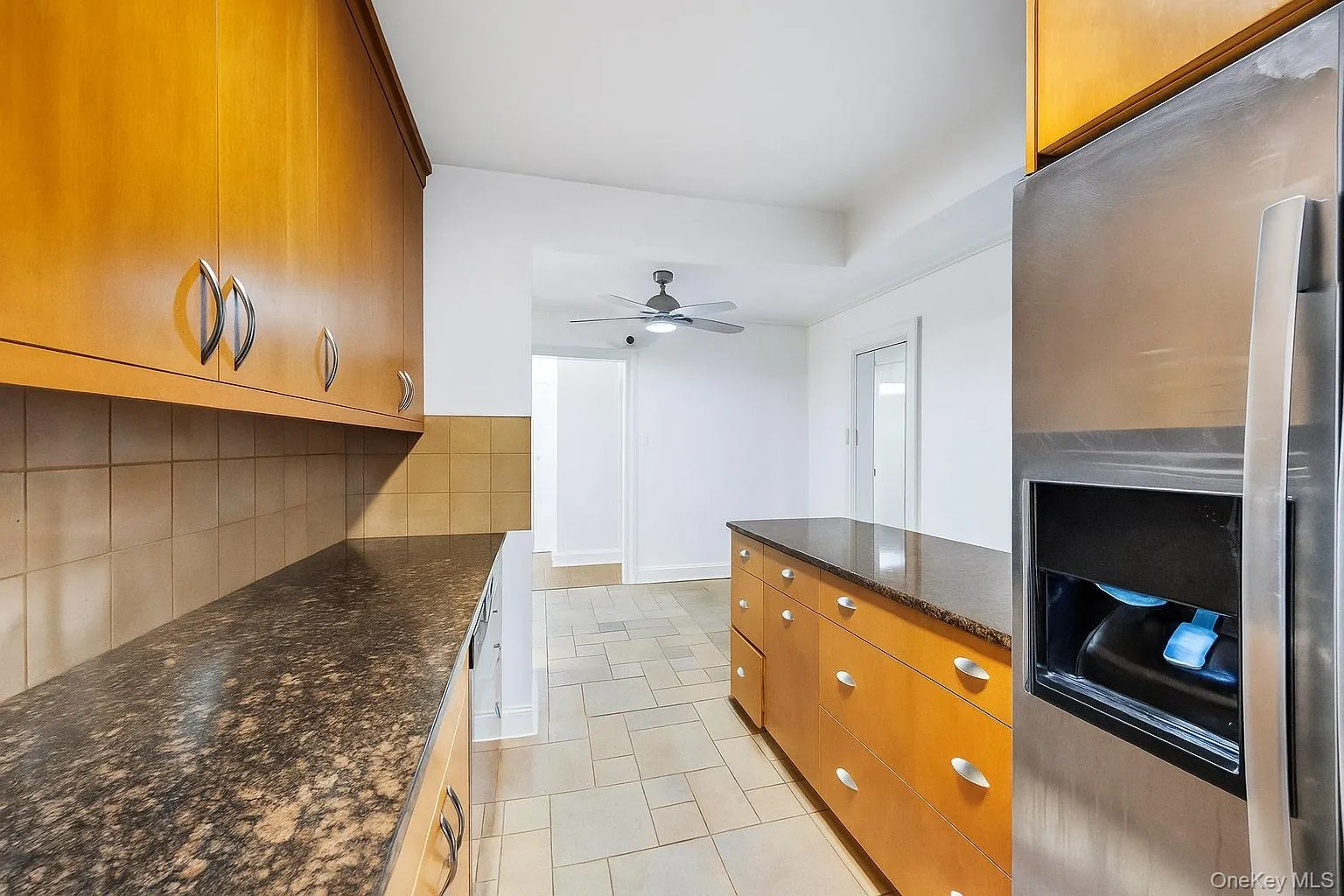 Kitchen with stainless steel fridge with ice dispenser, dark stone countertops, brown cabinets, decorative backsplash, and ceiling fan Kitchen with stainless steel fridge with ice dispenser, dark stone countertops, brown cabinets, decorative backsplash, and ceiling fan