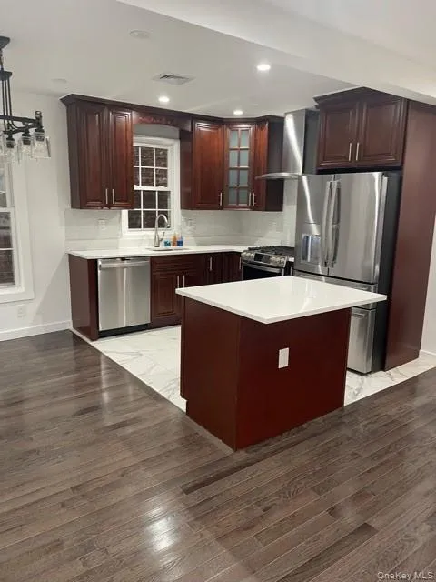 Kitchen with appliances with stainless steel finishes, light countertops, glass insert cabinets, wall chimney range hood, and a kitchen island Kitchen with appliances with stainless steel finishes, light countertops, glass insert cabinets, wall chimney range hood, and a kitchen island