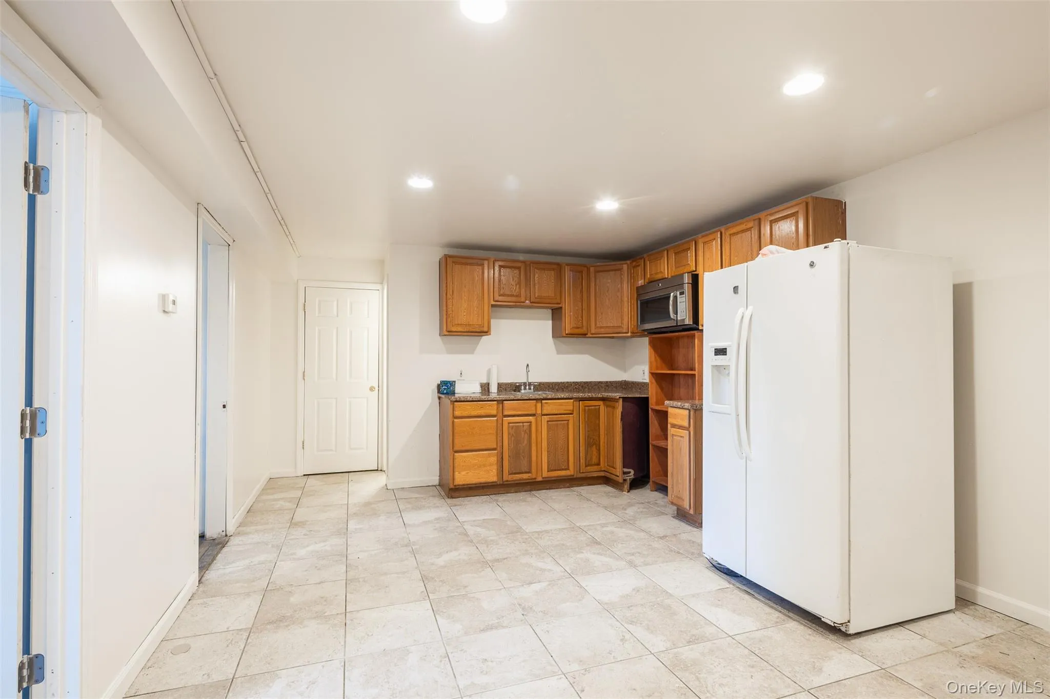 Kitchen featuring brown cabinets, white fridge with ice dispenser, recessed lighting, stainless steel microwave, and light tile patterned floors Kitchen featuring brown cabinets, white fridge with ice dispenser, recessed lighting, stainless steel microwave, and light tile patterned floors