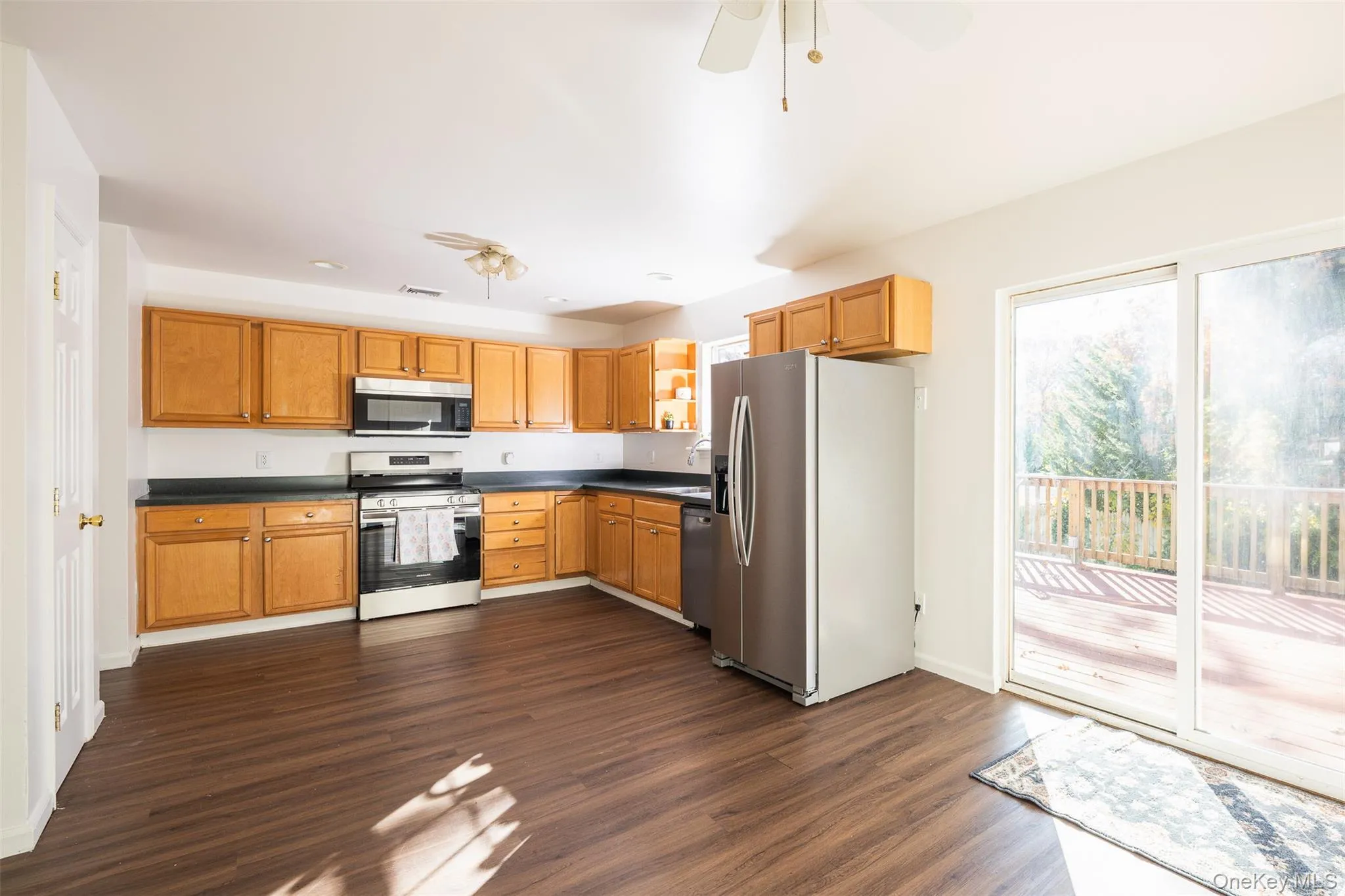 Kitchen featuring stainless steel appliances, dark countertops, dark wood finished floors, brown cabinetry, and a ceiling fan Kitchen featuring stainless steel appliances, dark countertops, dark wood finished floors, brown cabinetry, and a ceiling fan