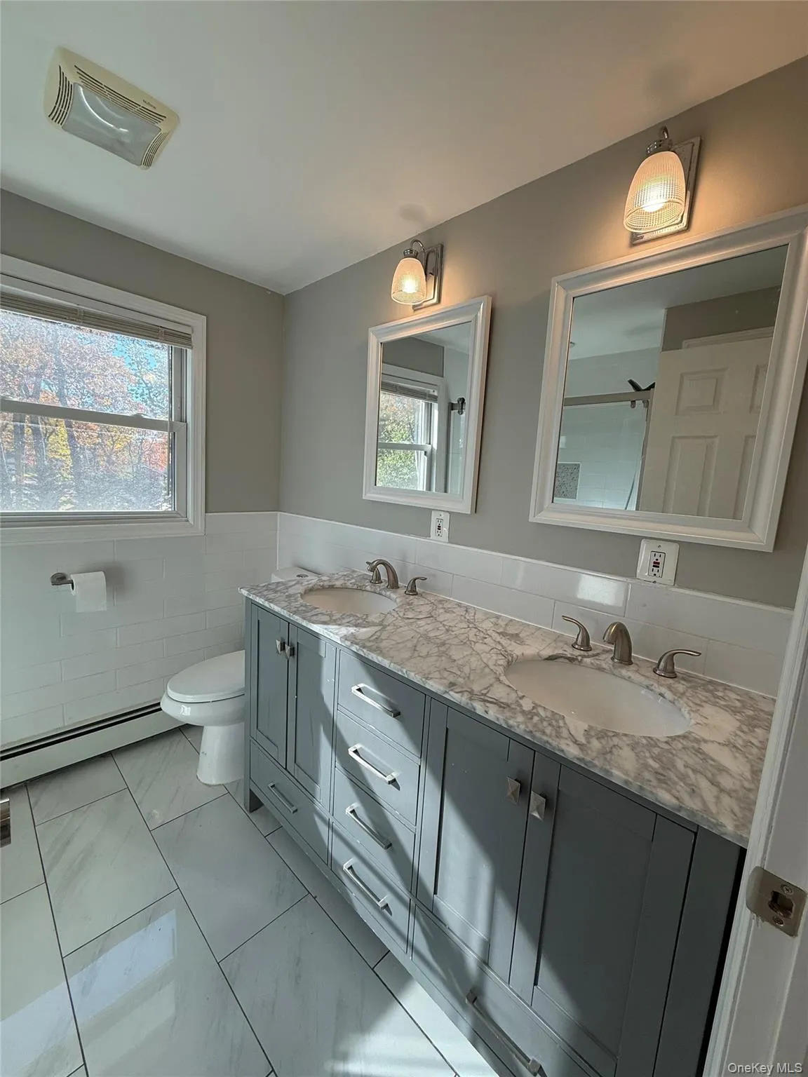 Bathroom featuring double vanity, tile walls, a baseboard heating unit, light marble finish flooring, and a wainscoted wall Bathroom featuring double vanity, tile walls, a baseboard heating unit, light marble finish flooring, and a wainscoted wall