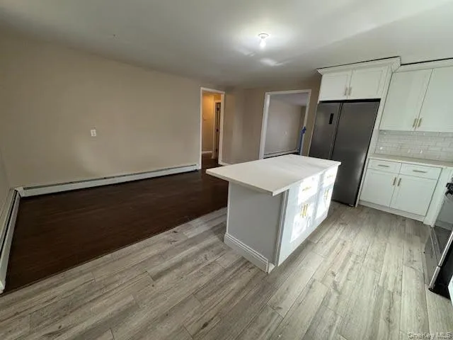 Kitchen with white cabinets, decorative backsplash, a center island, freestanding refrigerator, a baseboard radiator, kitchen window and an island for extra counter top area. Kitchen with white cabinets, decorative backsplash, a center island, freestanding refrigerator, a baseboard radiator, kitchen window and an island for extra counter top area.